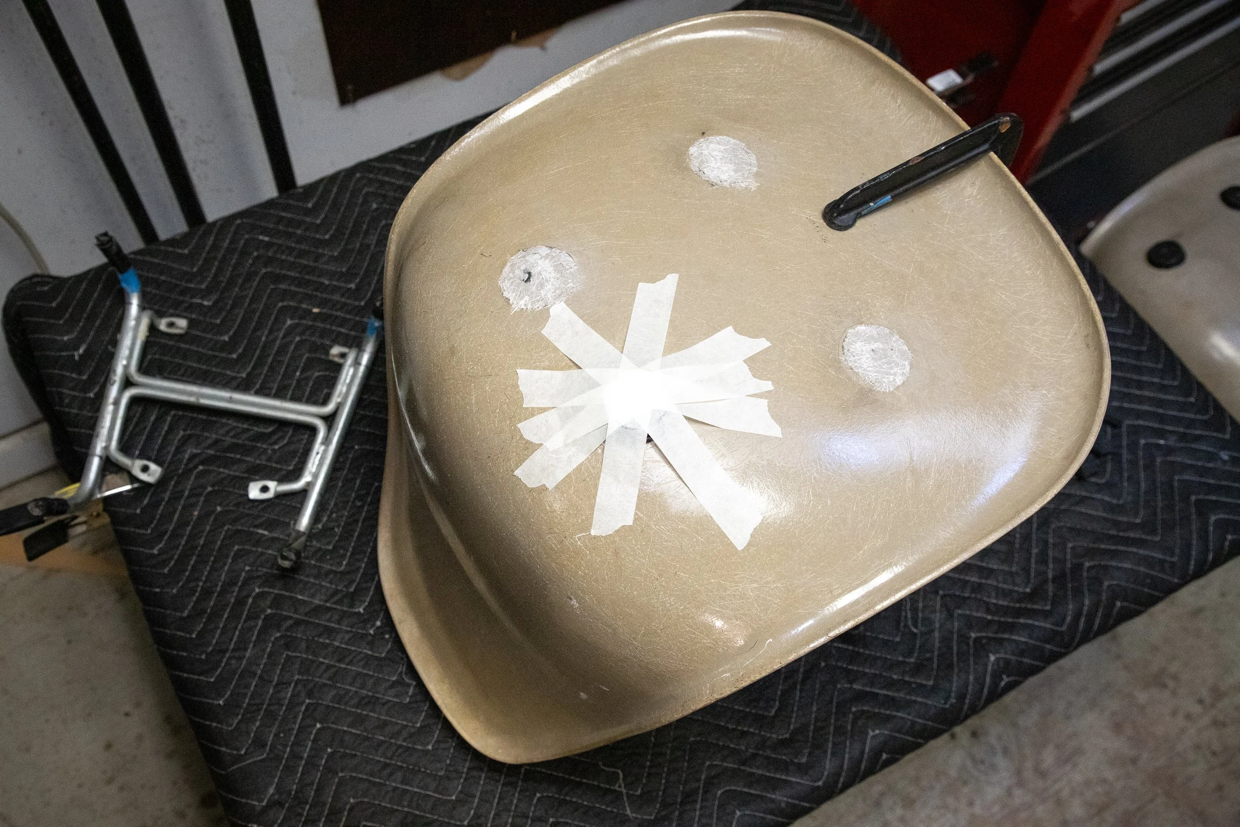 A beige plastic school chair with four white tape marks on the seat, placed on a black quilted blanket or pad. In the background, a headless white plastic chair and some tools or objects are visible.