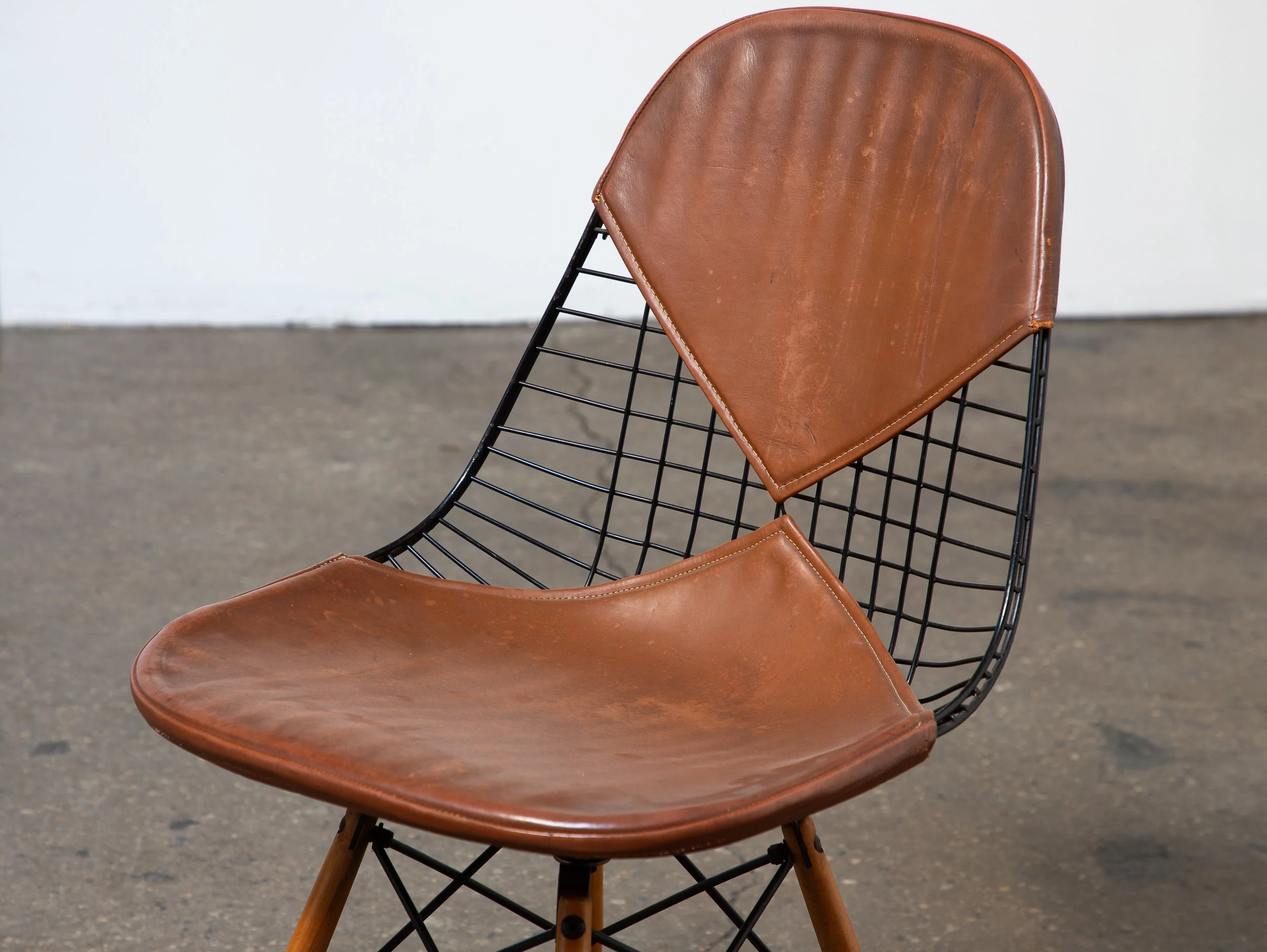 A mid-century modern style chair with a leather seat and backrest, and black metal wireframe sides and wooden legs on a gray concrete floor against a plain white wall.