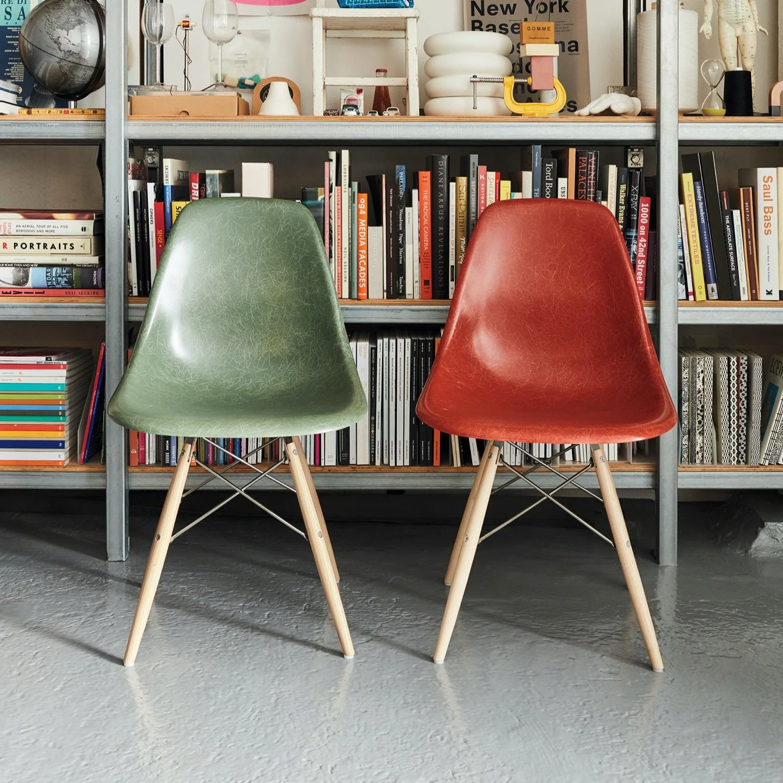Two vintage-style chairs, one green and one red, with wooden legs and metal supports, are placed in front of a metal bookshelf filled with books, decorative objects, and framed pictures.