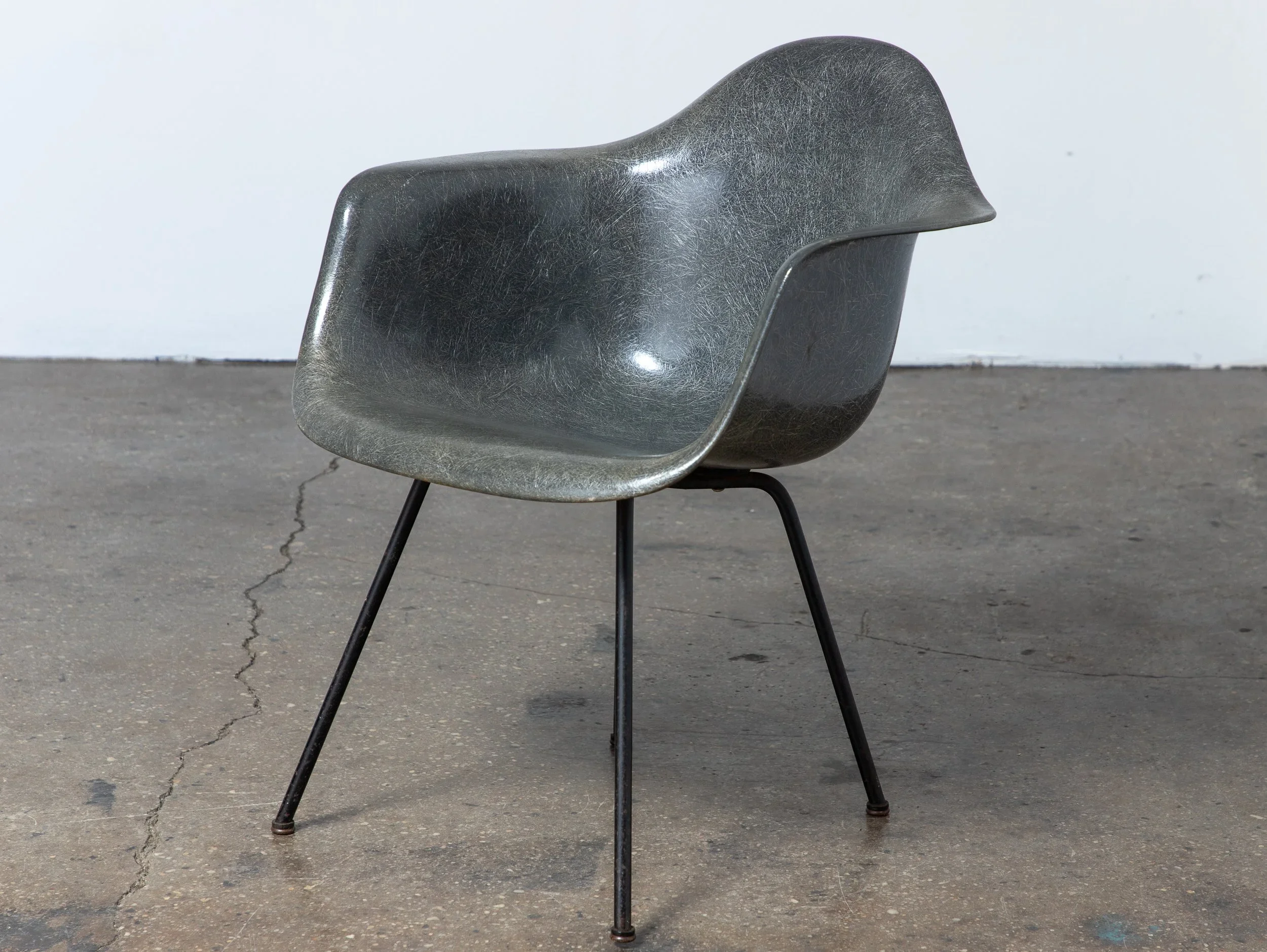 A modern black plastic chair with metal legs is placed on a concrete surface outside, with a plain light-colored wall in the background.