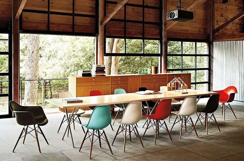 Modern conference room with a long white table surrounded by colorful chairs, large windows showing trees outside, and wooden paneling on the ceiling.