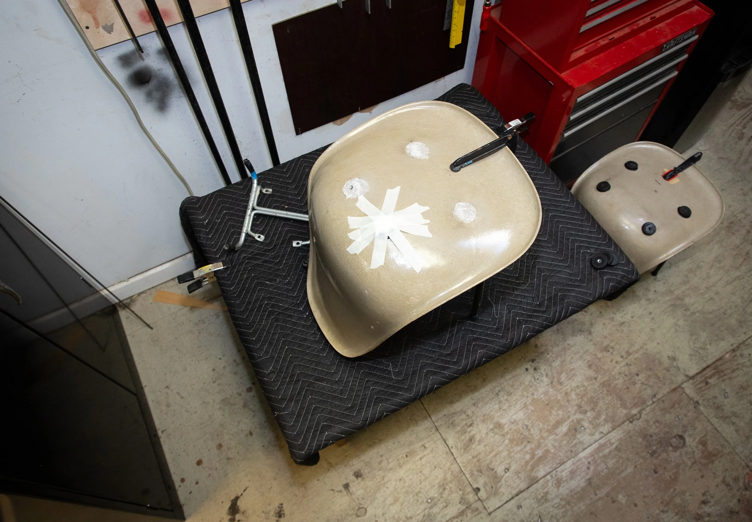 A beige plastic chair with a black padded seat placed on a black padded cloth on the floor in a workshop or garage setting. There are tape and some tools on the chair, and in the background, a red tool chest, a white wall with black marks, and some tools hanging on the wall.