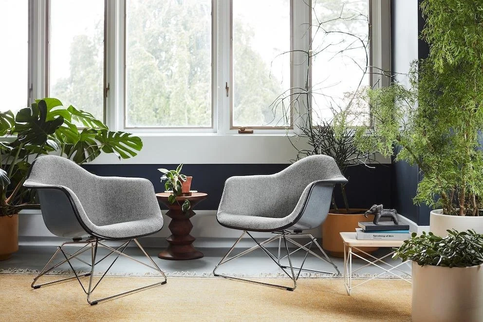A modern indoor sitting area with two gray armchairs, a small wooden side table, a stack of books with a small sculpture, and various large green plants near a window.