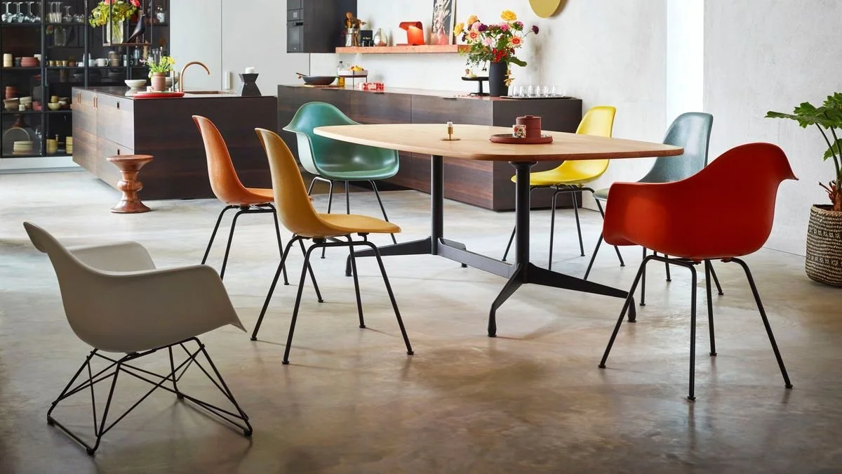 Modern dining area with a round table and colorful chairs, a dark kitchen island in the background, and a vase with flowers on a side buffet.