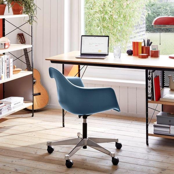 Home office workspace with a blue swivel chair, wooden desk, laptop, books, and a guitar in a bright room with large window and green outdoor view.