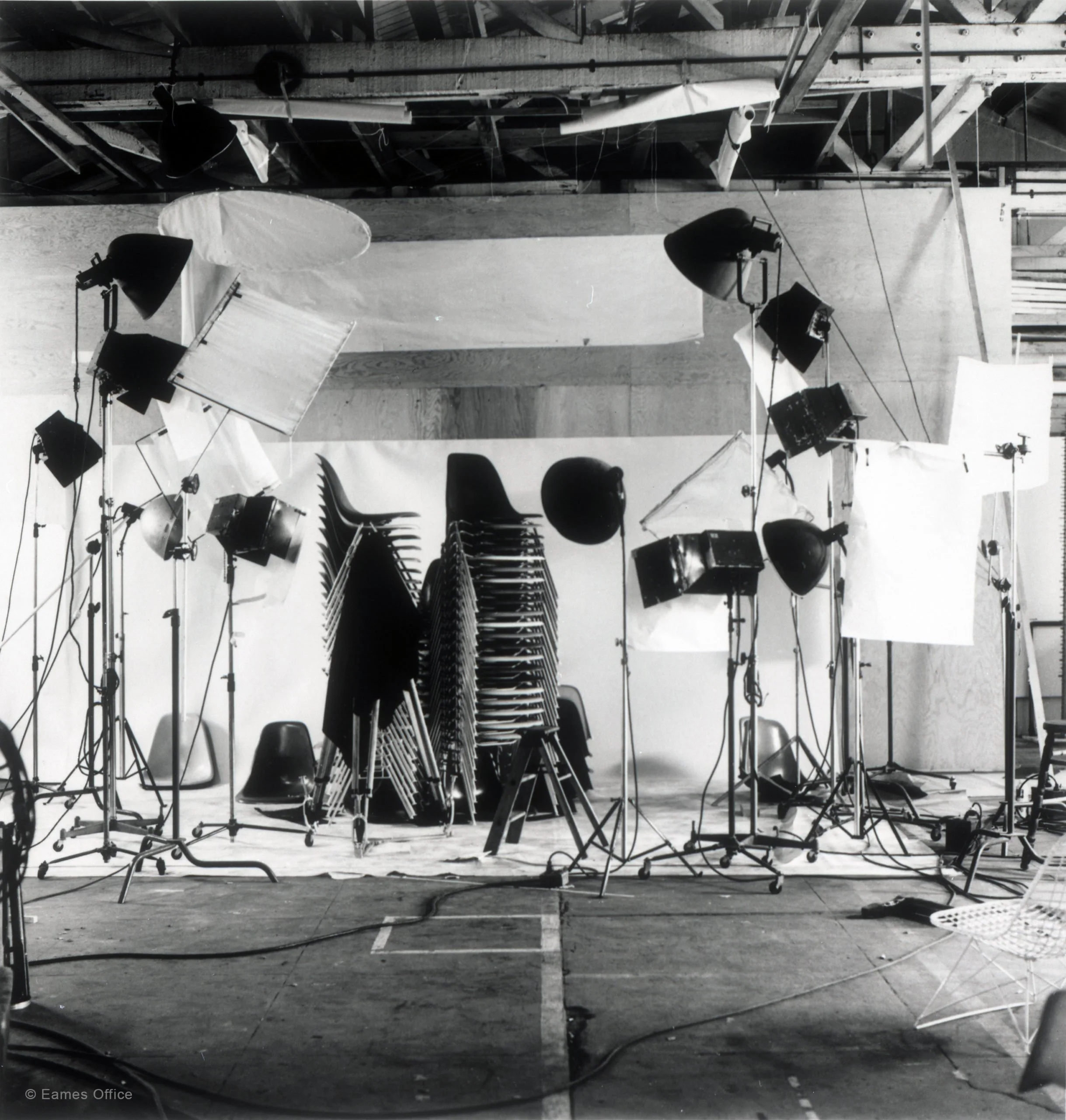A black and white photo of a photography studio setup showing stacked chairs, studio lighting with softbox diffusers, and various equipment in a large open space with a wooden ceiling.