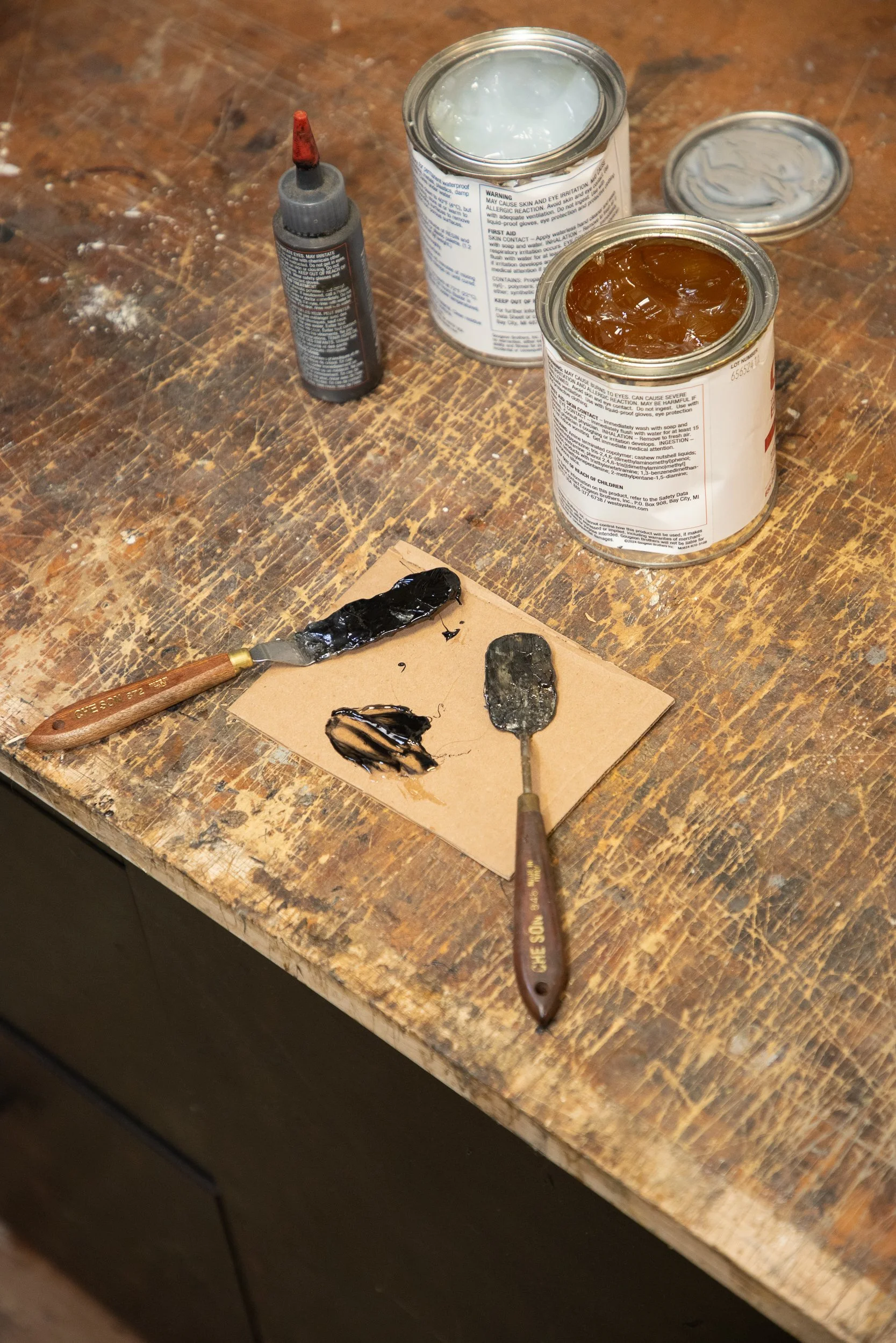 Open cans of black and brown art mediums, a small tube of liquid black paint, and two palette knives with black paint on them, on a wooden table.