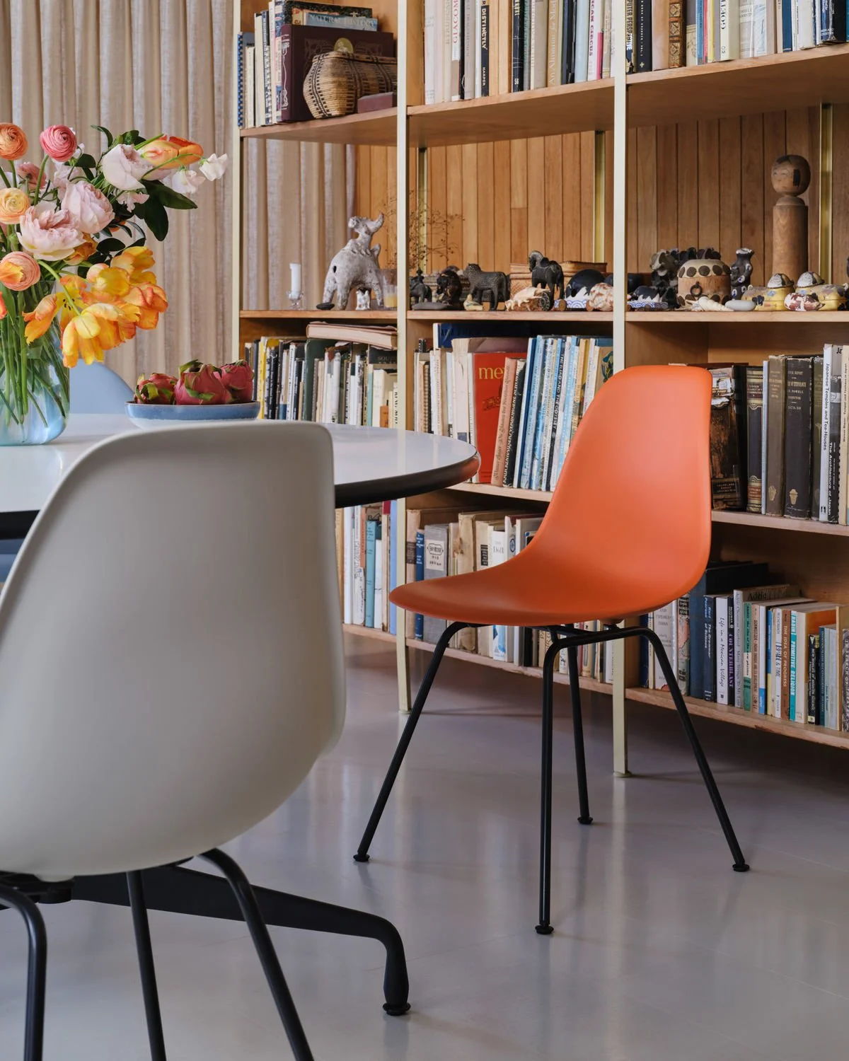 Interior of a room with a white round table, a white chair, and an orange chair, with a wooden bookshelf filled with books and decorative objects in the background, and a vase of colorful flowers on the table.