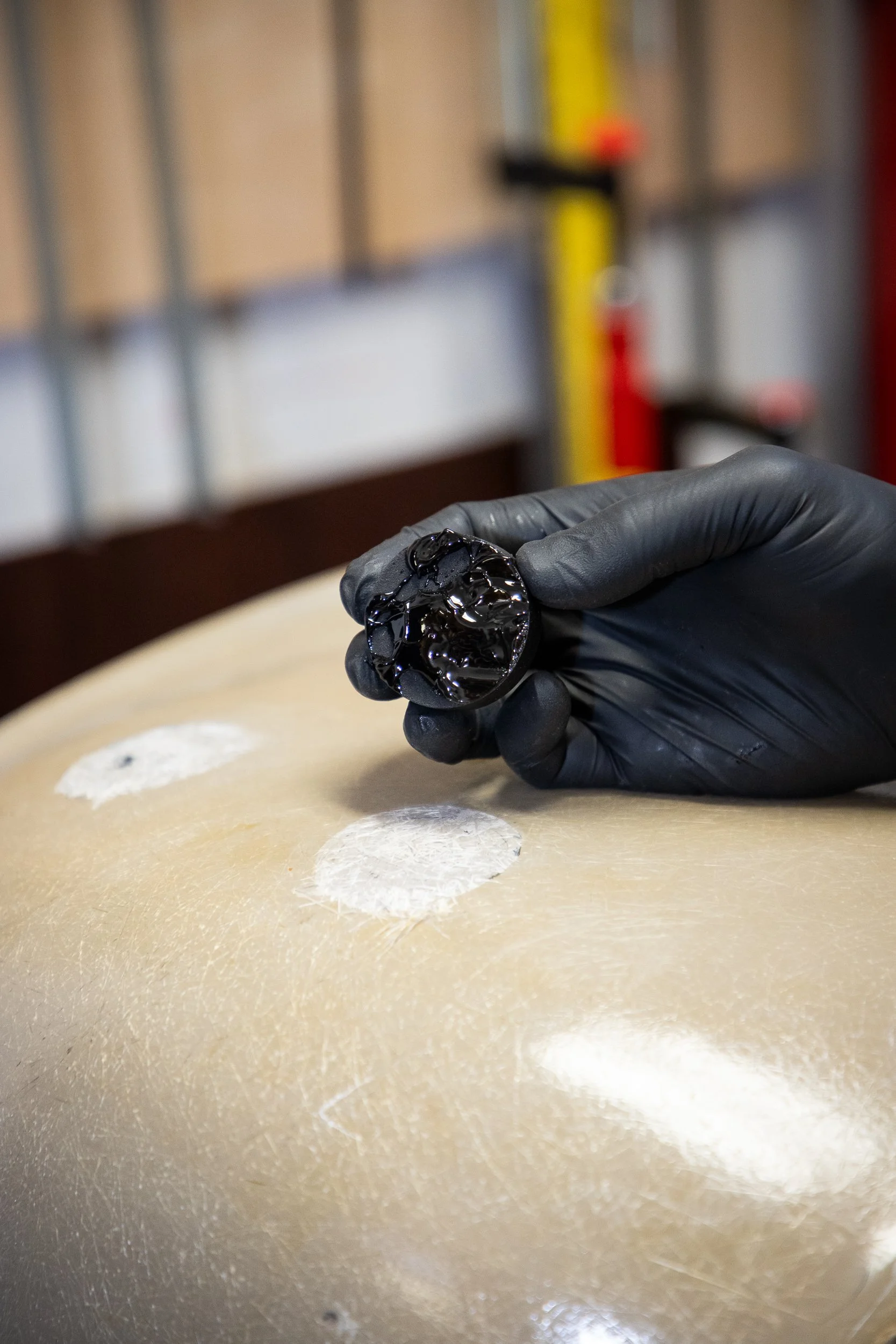 A person wearing black gloves holding a small, shiny, black object, possibly a piece of jewelry, over a light-colored surface with white circular spots, with a blurred background of a workshop or industrial setting.