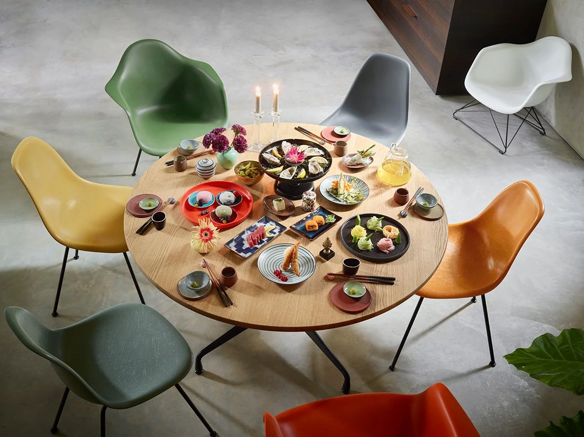 A round wooden dining table set with colorful chairs, decorated with various plates of sushi, a sake set, flowers, and candlesticks, in a modern dining room.