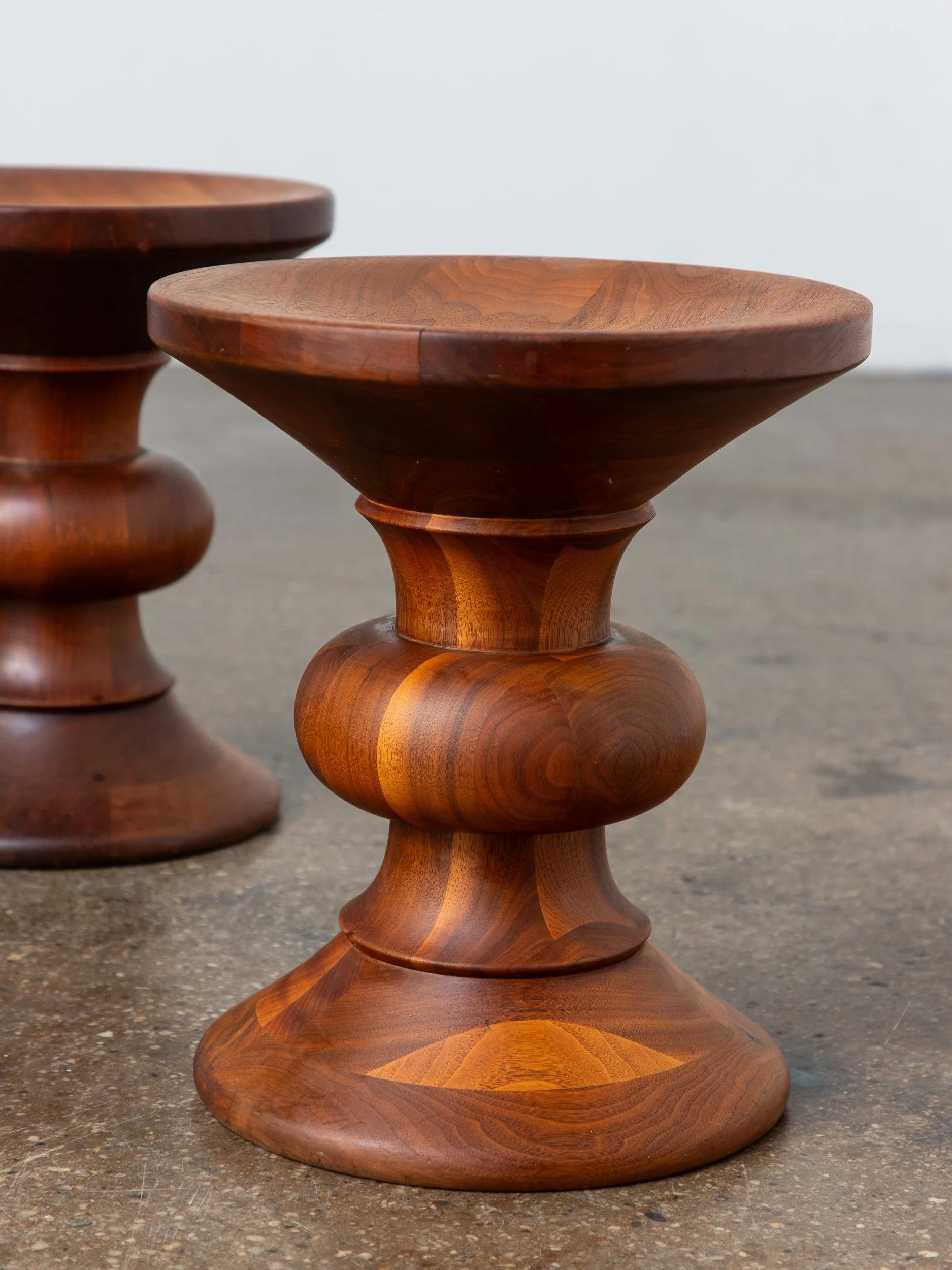 Close-up of two polished wooden stools with rounded bases and wide, shallow tops, placed on a concrete floor against a plain white wall.