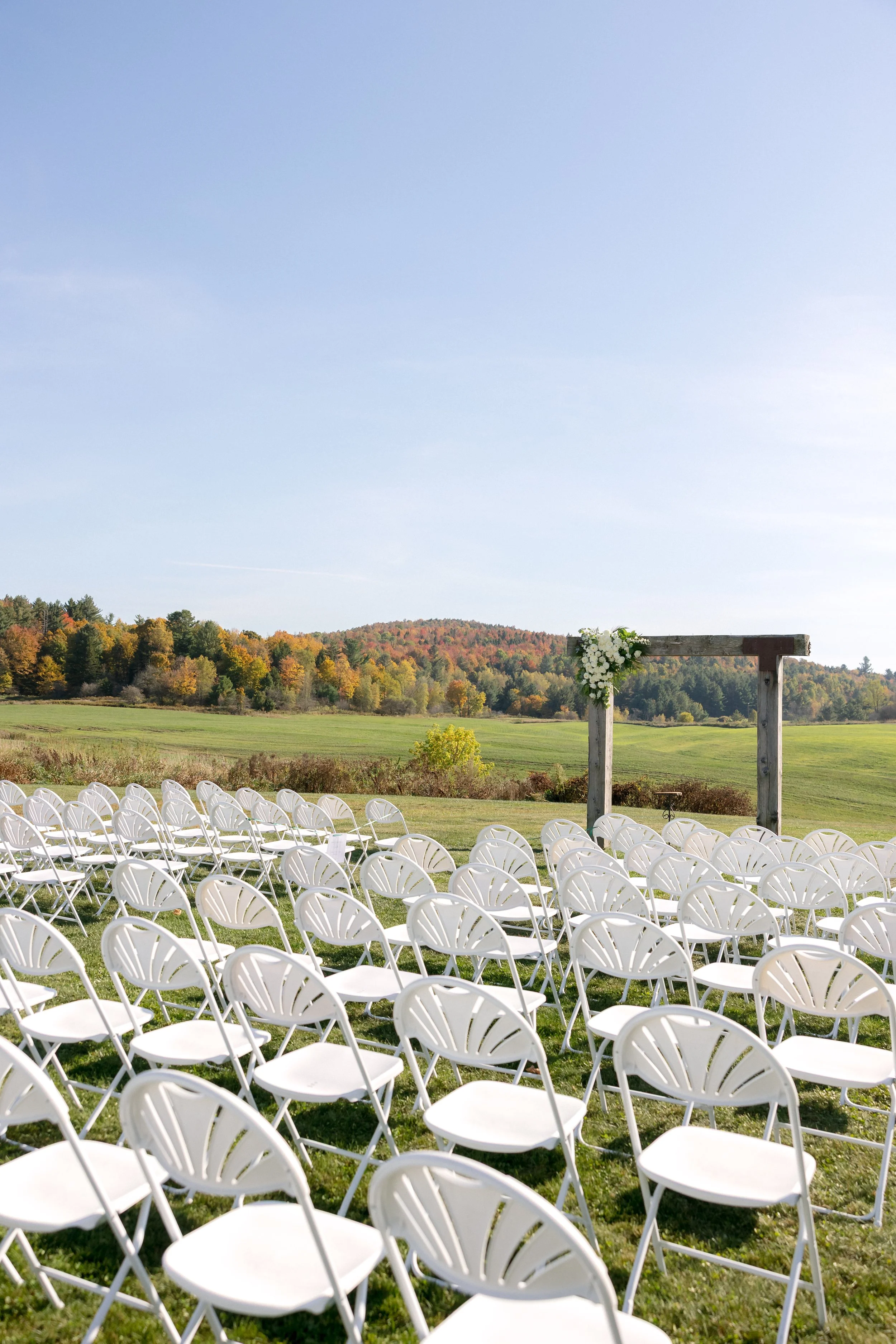 Outdoor meadow ceremony setup with white chairs and a wooden arch overlooking the Vermont hills at Swiss & Bloom Farm.