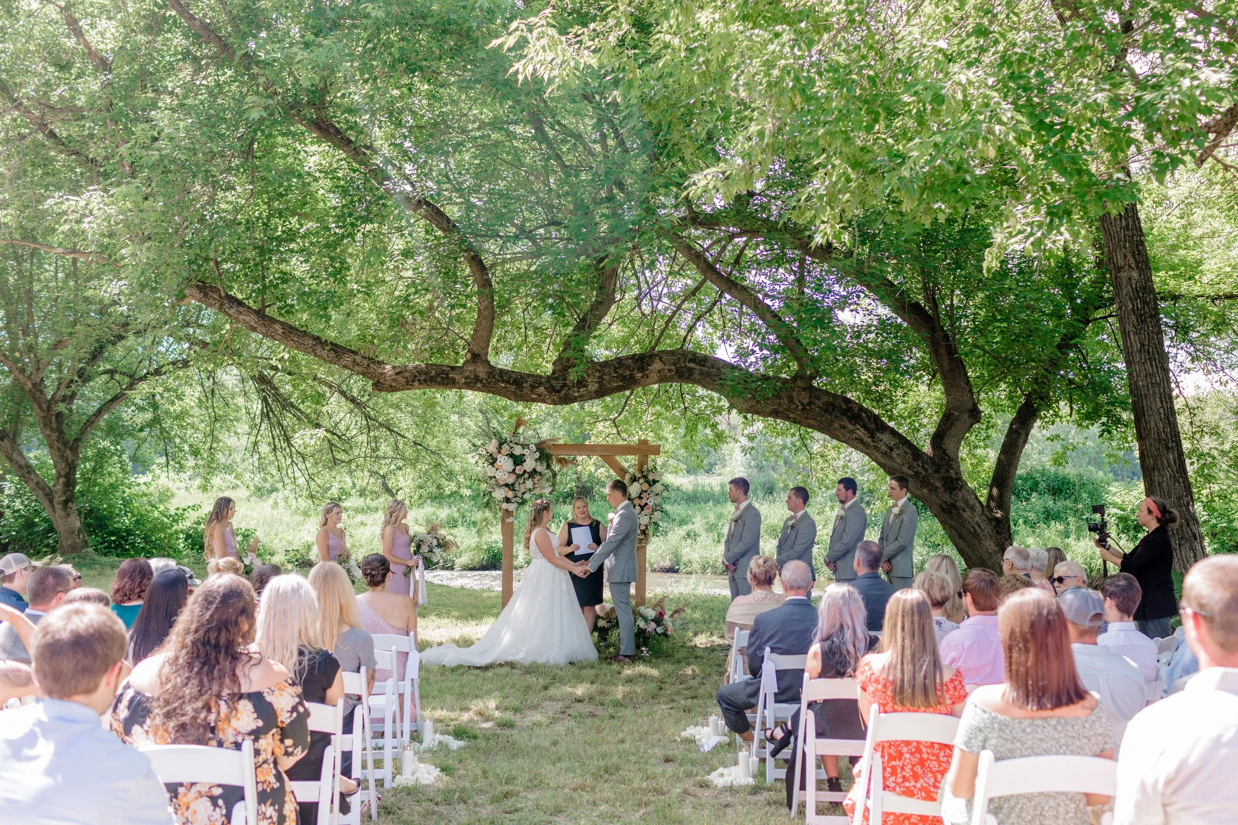 Bride and groom exchanging vows beneath a tree canopy with guests seated outdoors at Swiss & Bloom Farm.