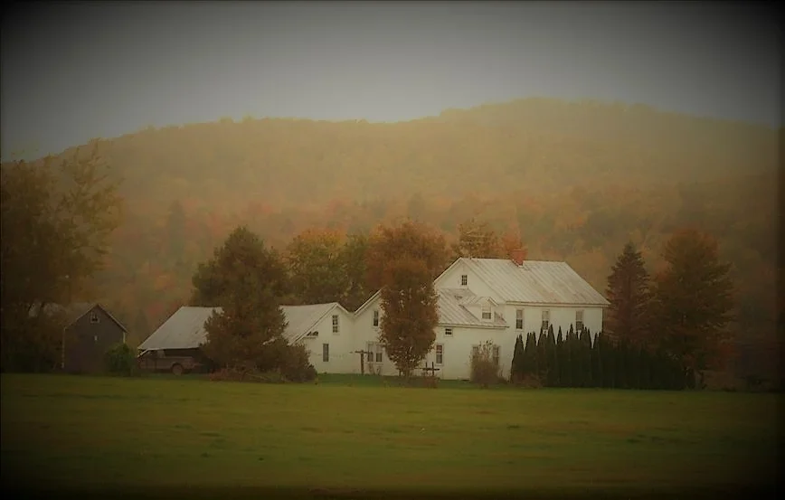 The historic 1885 Swiss & Bloom Farmstead in autumn, with the farmhouse and barn framed by colorful Vermont foliage.