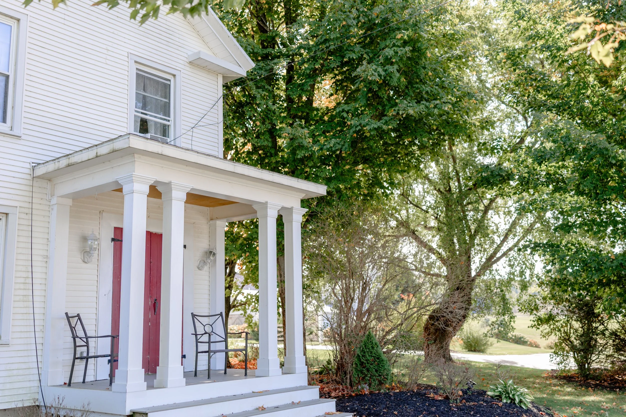 The white farmhouse at Swiss & Bloom Farm, offering on‑site wedding lodging with a shaded porch and surrounding trees.