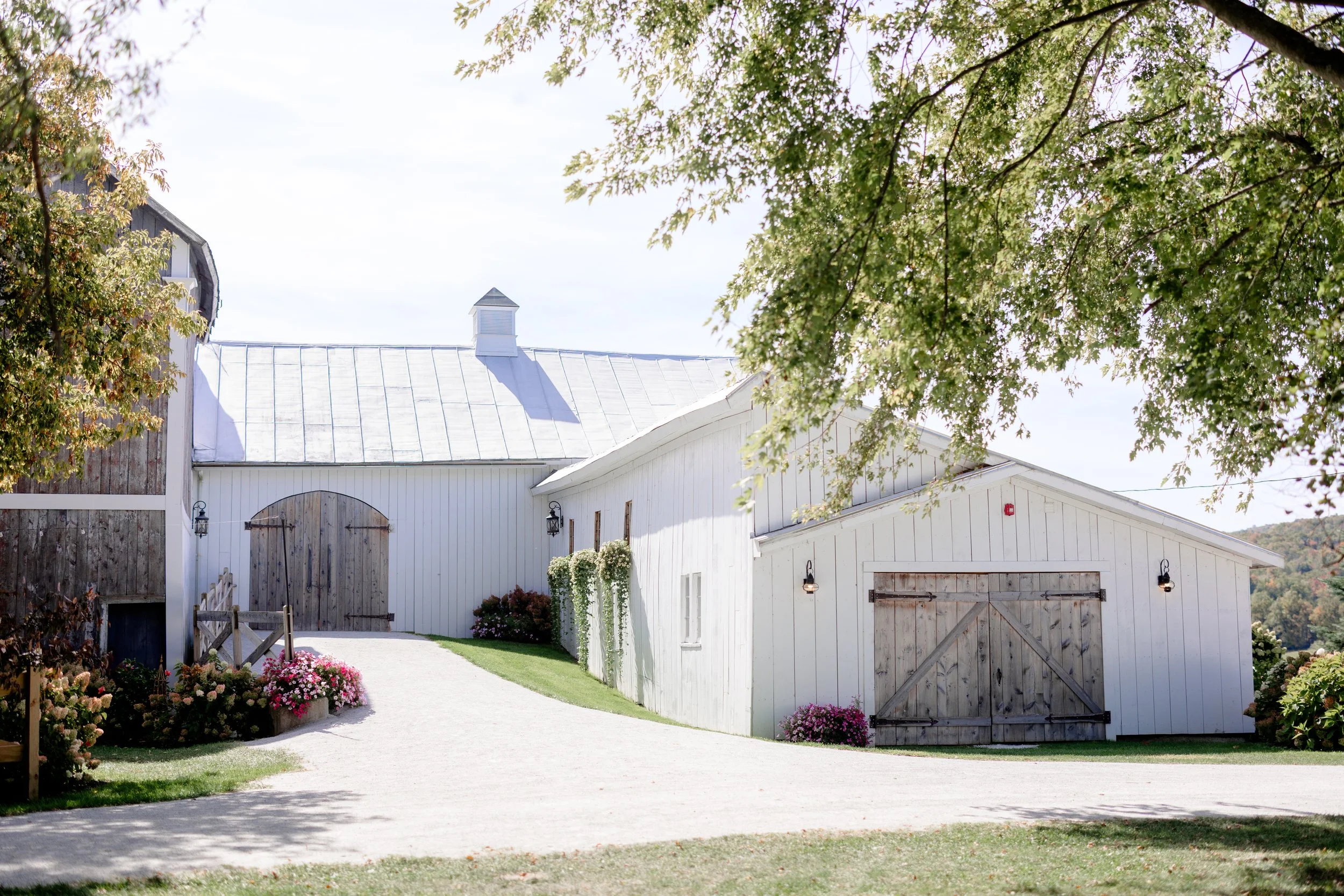 Side entrance view of the Wedding Barn at Swiss & Bloom Farm with large wooden barn doors, gravel drive, and surrounding trees.