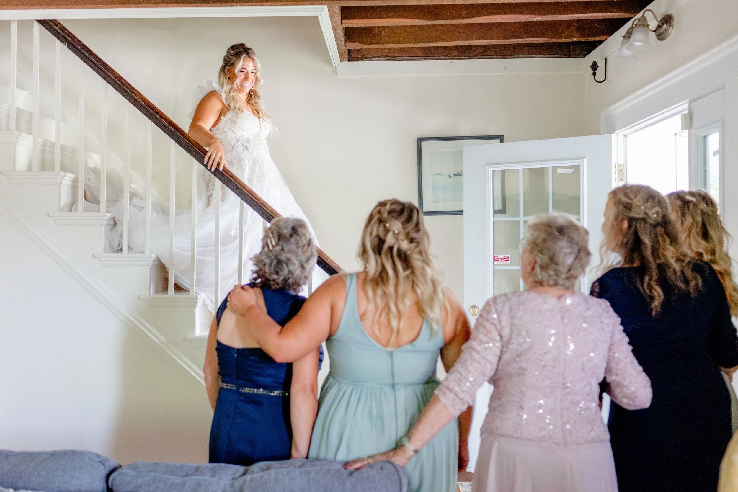 Bride standing on the farmhouse staircase as her bridesmaids look up during wedding‑day preparations at Swiss & Bloom Farm.
