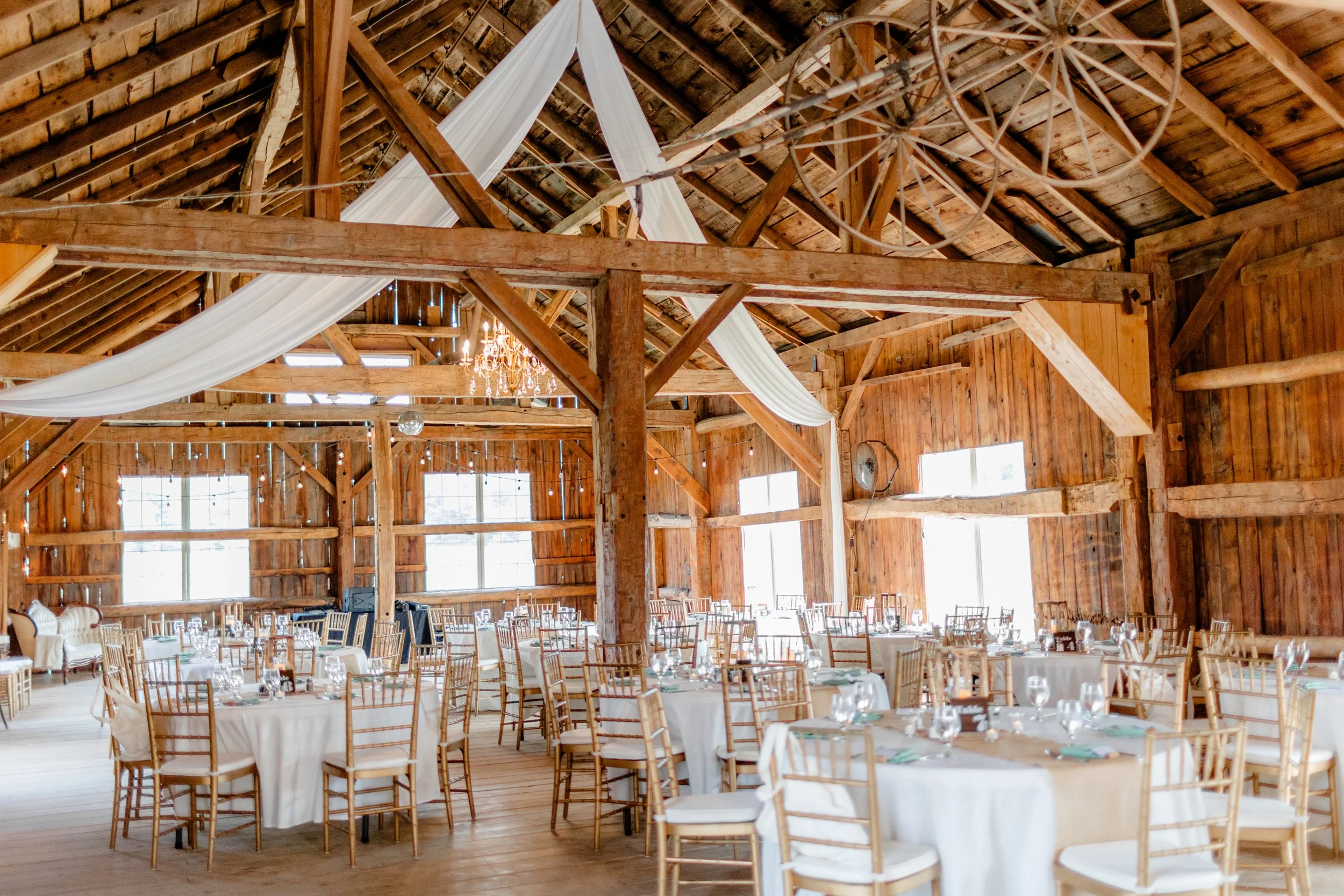 Interior of the restored wedding barn at Swiss & Bloom Farm set for a reception with wooden beams, draped fabric, and warm lighting.