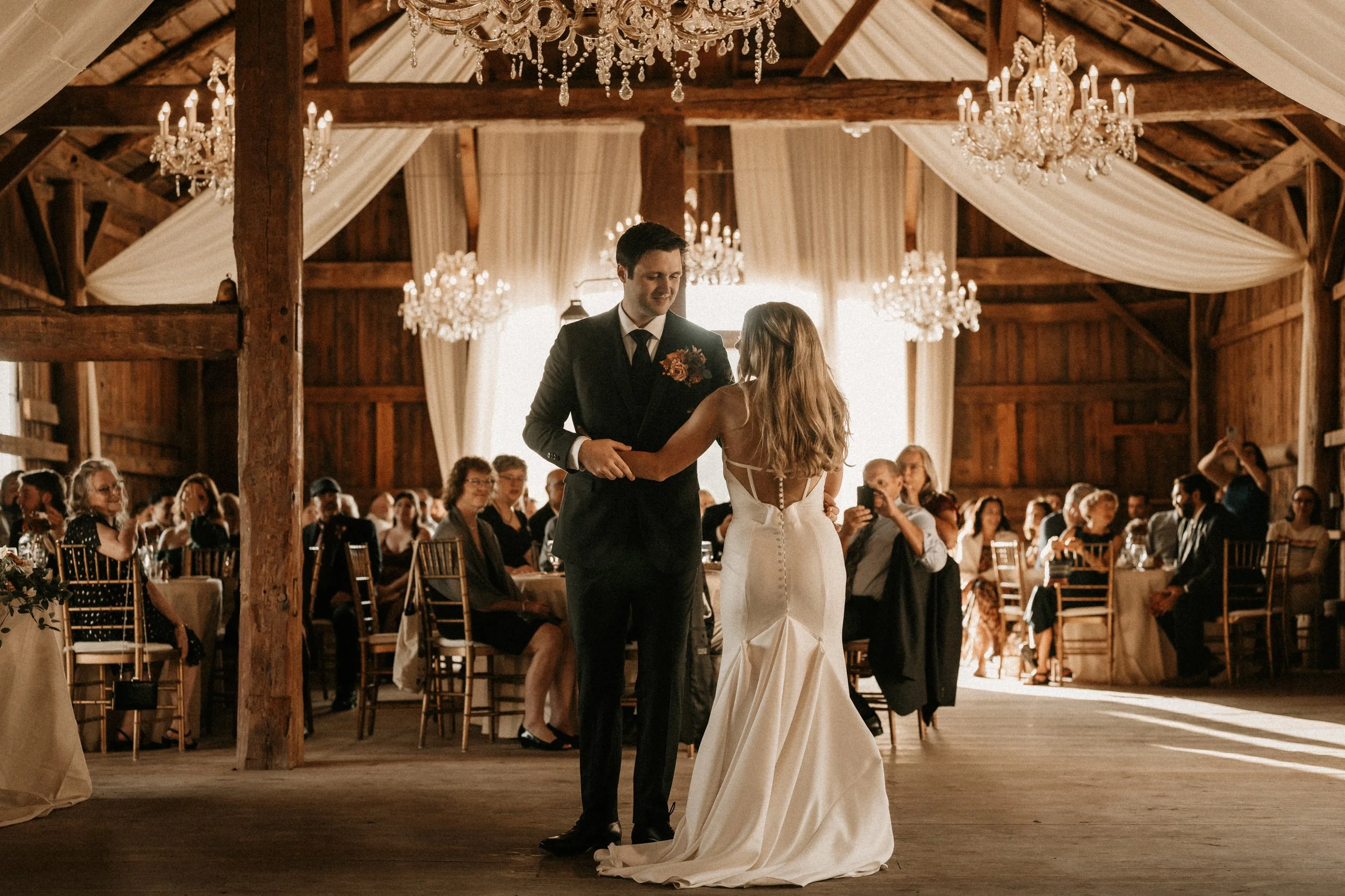 Bride and groom sharing a dance beneath chandeliers and soft draping inside the Wedding Barn at Swiss & Bloom Farm.
