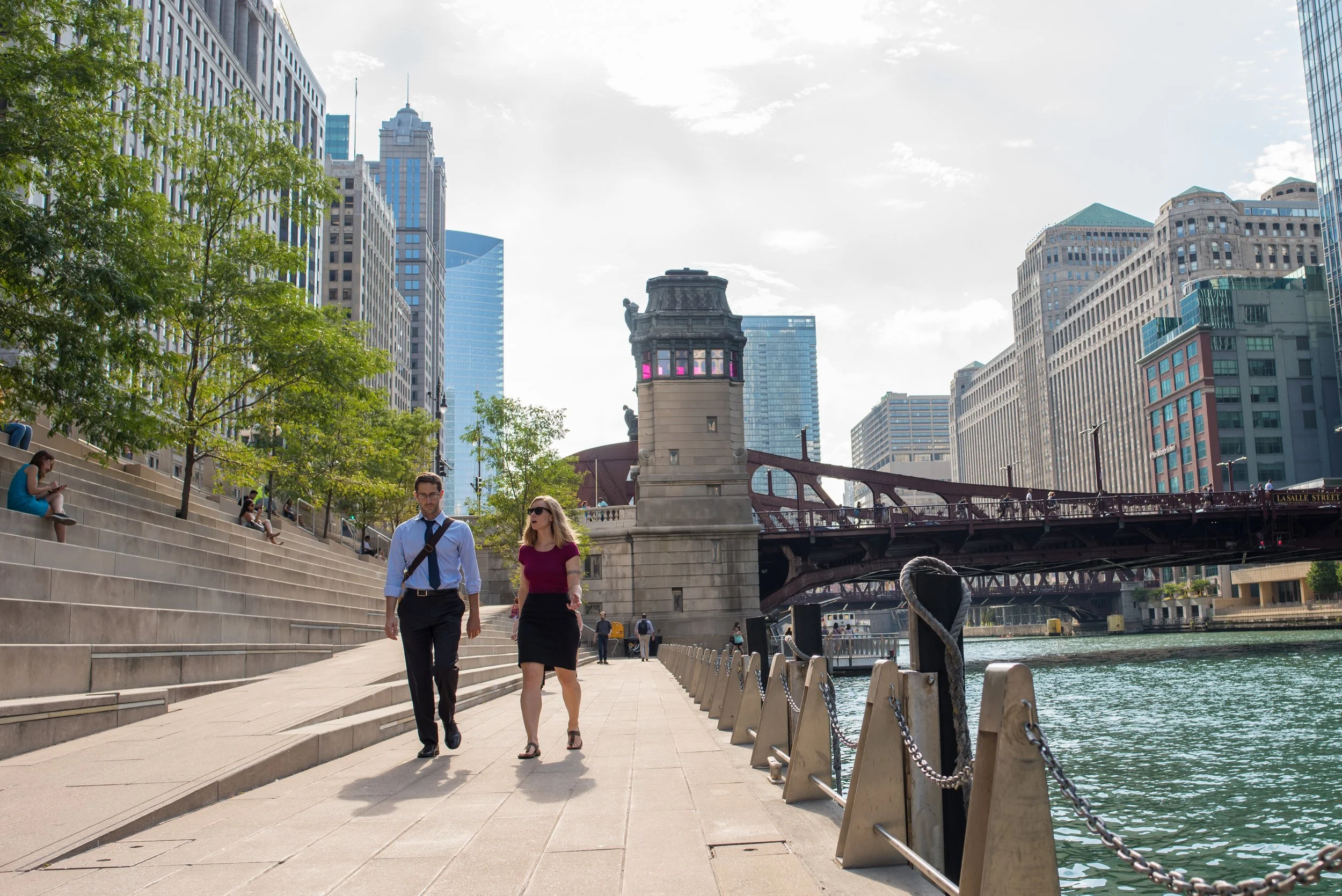 People walking along the riverwalk in a city with high-rise buildings, a bridge, and trees.