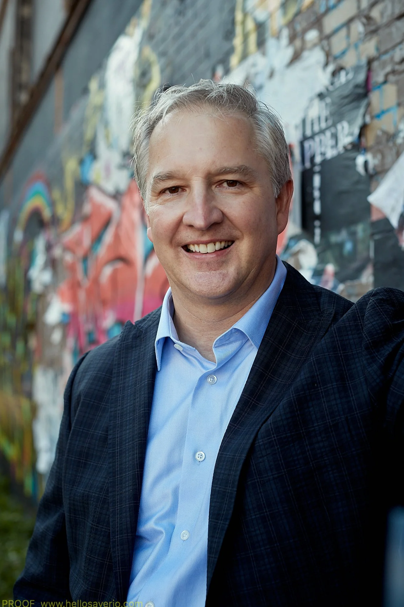 A middle-aged man with gray hair smiling outdoors in front of a colorful graffiti wall.
