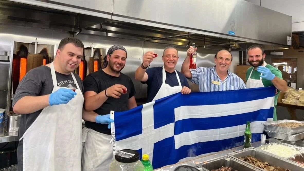Six men standing in a restaurant kitchen holding a Greek flag, smiling and raising small drinks.
