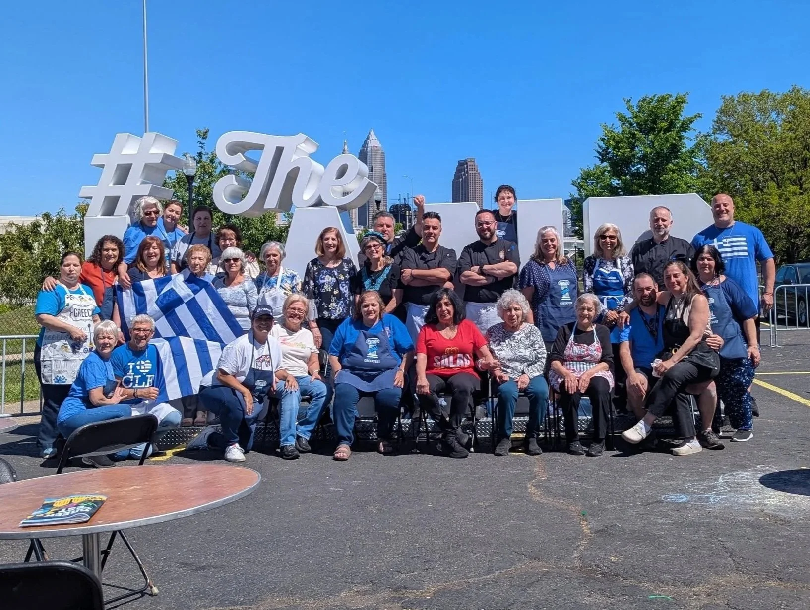 Group of people posing outdoors in front of a large sign that says '#The' with a city skyline in the background, some holding Greek flags, all wearing casual clothing.