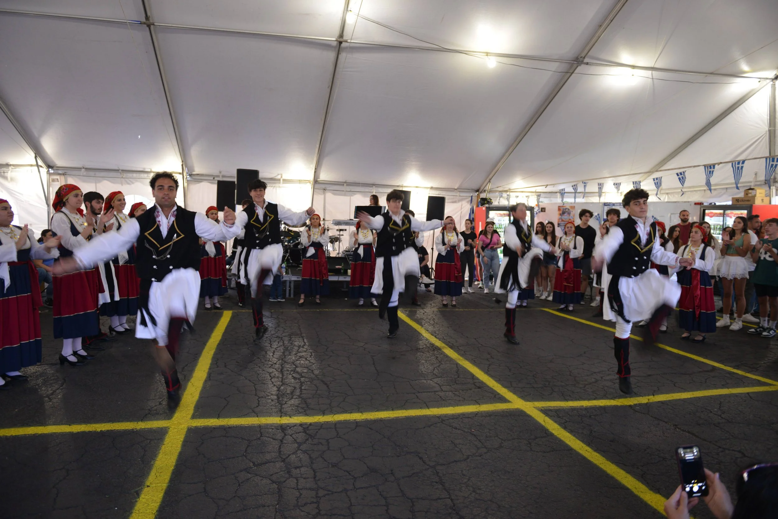Greek dancers performing a traditional dance inside a large tent, wearing traditional costumes, with an audience watching.