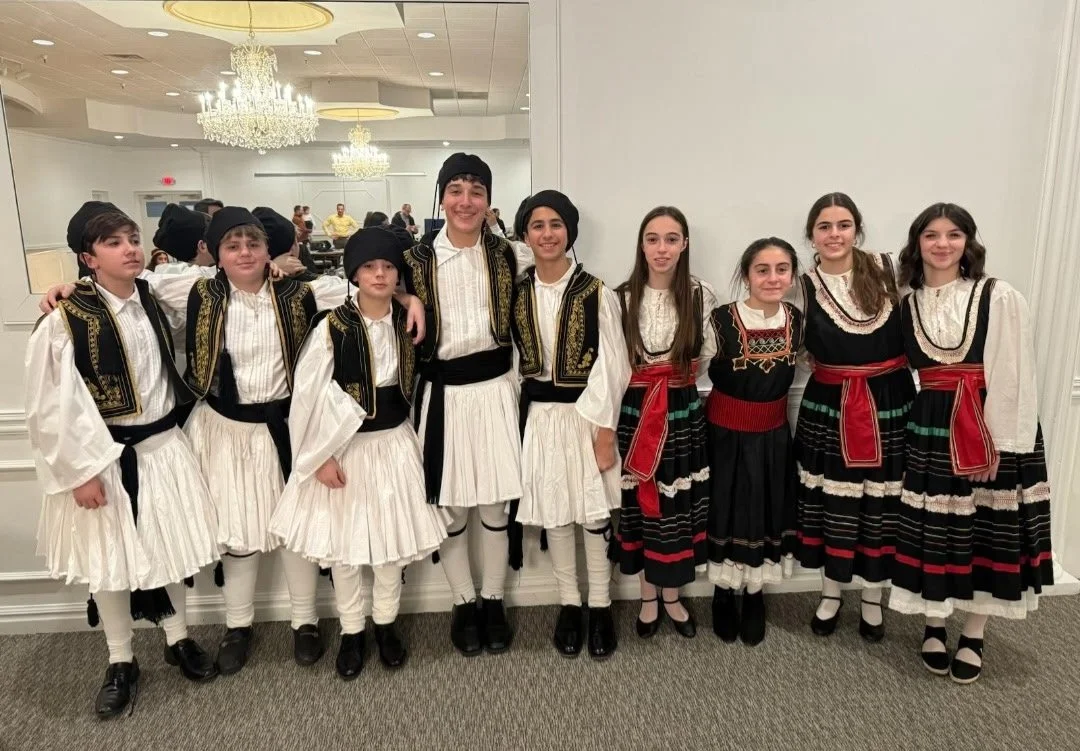 Group of children in traditional Greek costumes posing inside a banquet hall with chandeliers.