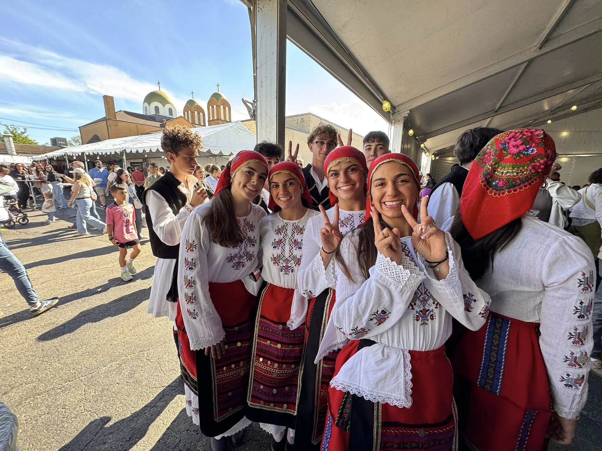 Group of young women and men dressed in traditional Bulgarian folk costumes at an outdoor festival.