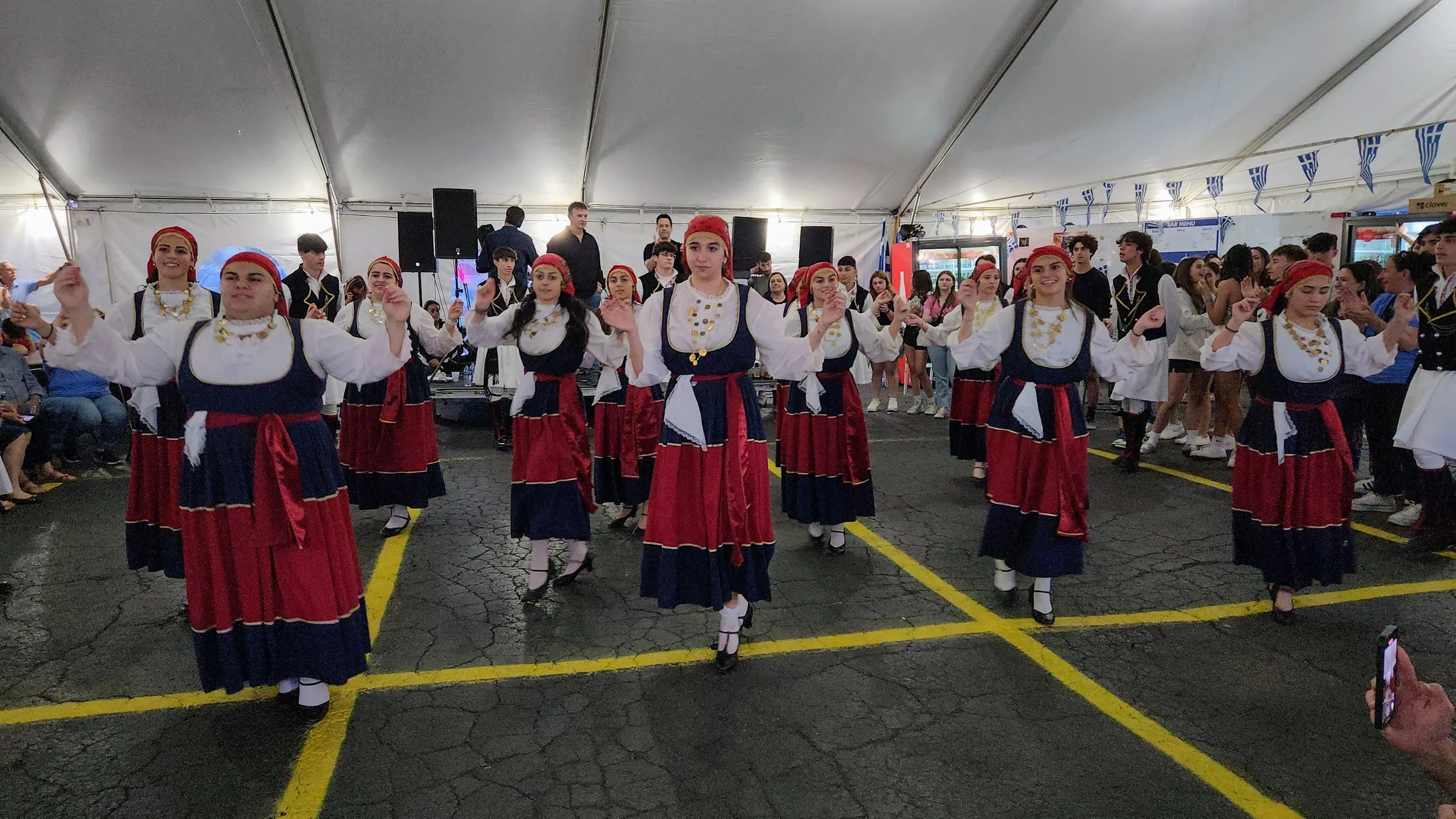 Group of women and men in traditional Greek costumes performing a dance inside a large tent at a cultural event.
