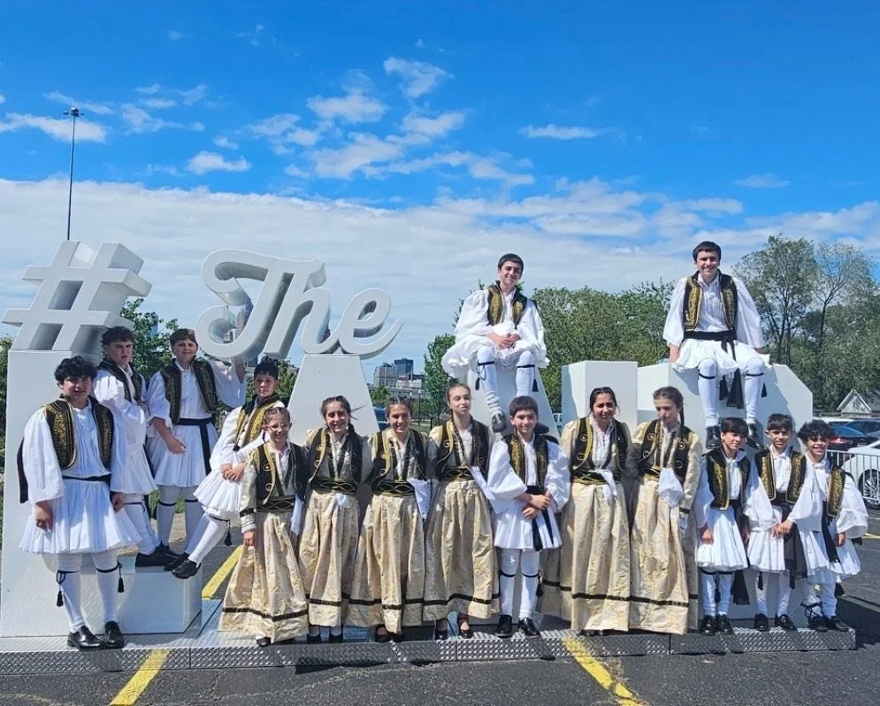 Group of children and teens in traditional Greek costumes posing in front of large white hashtag and text '#The' sign during daytime, with clear skies and trees in the background.
