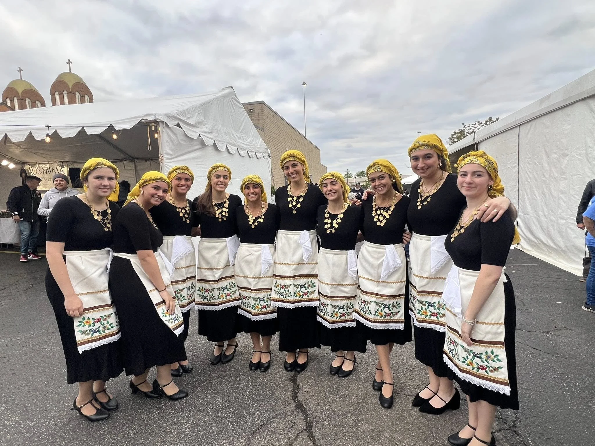 Group of eleven women dressed in traditional folk clothing, black tops, cream aprons with embroidered floral patterns, yellow headscarves, and black shoes, standing outdoors at a cultural event.