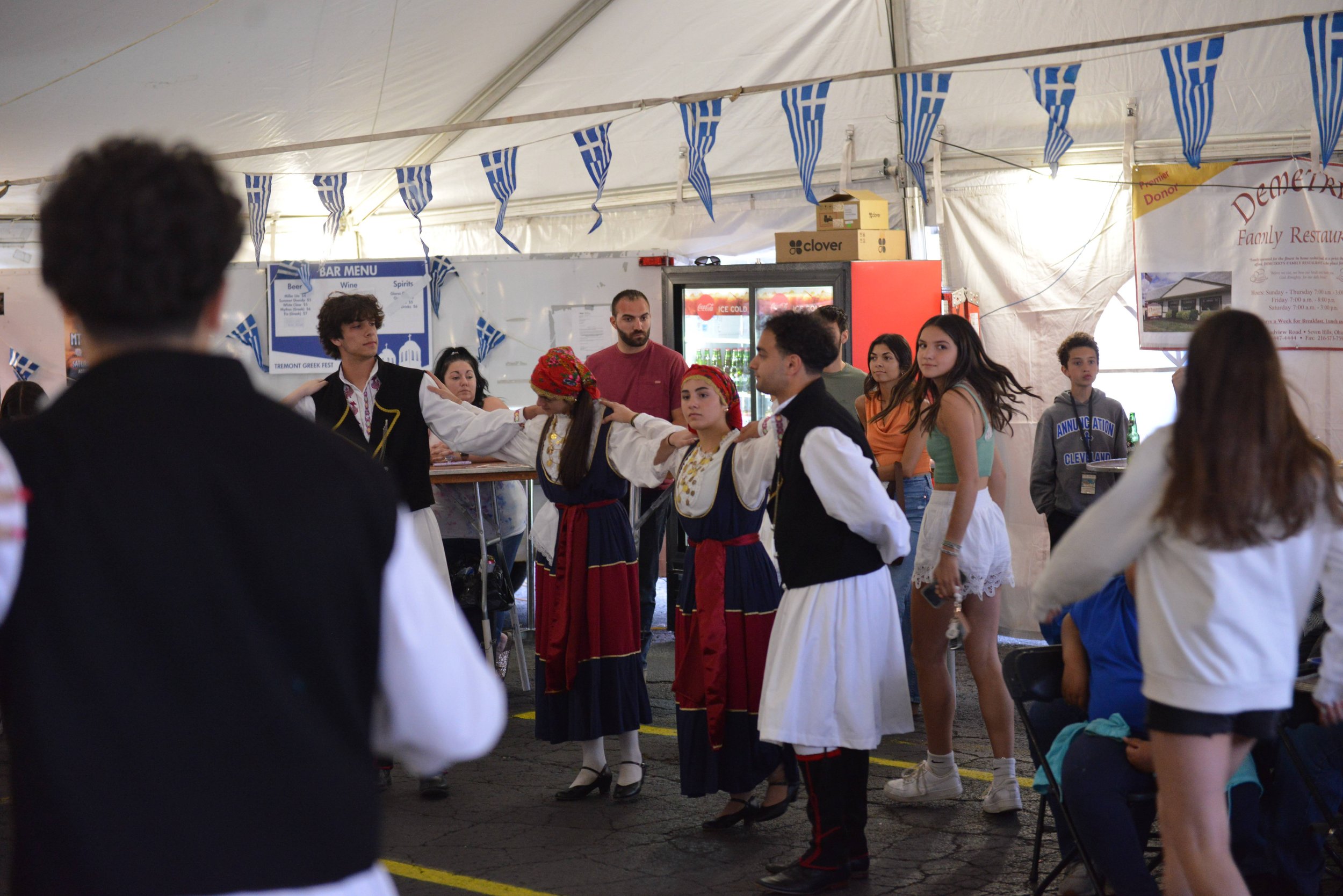 People performing a traditional Greek dance in a tent decorated with Greek flags during a cultural festival.