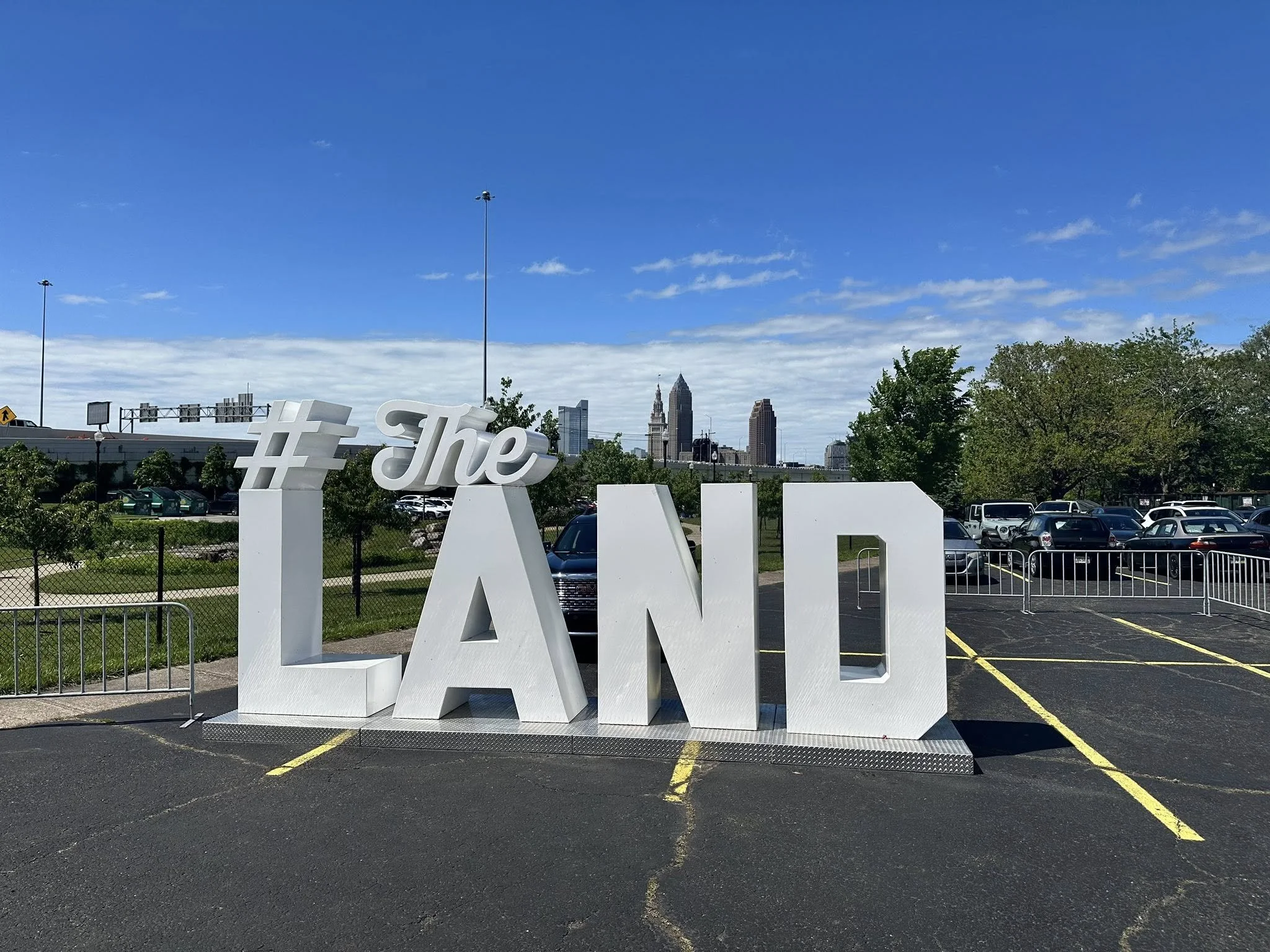 Large white 3D letters spelling '#The LAND' on a parking lot with city skyline in the background under a partly cloudy blue sky.