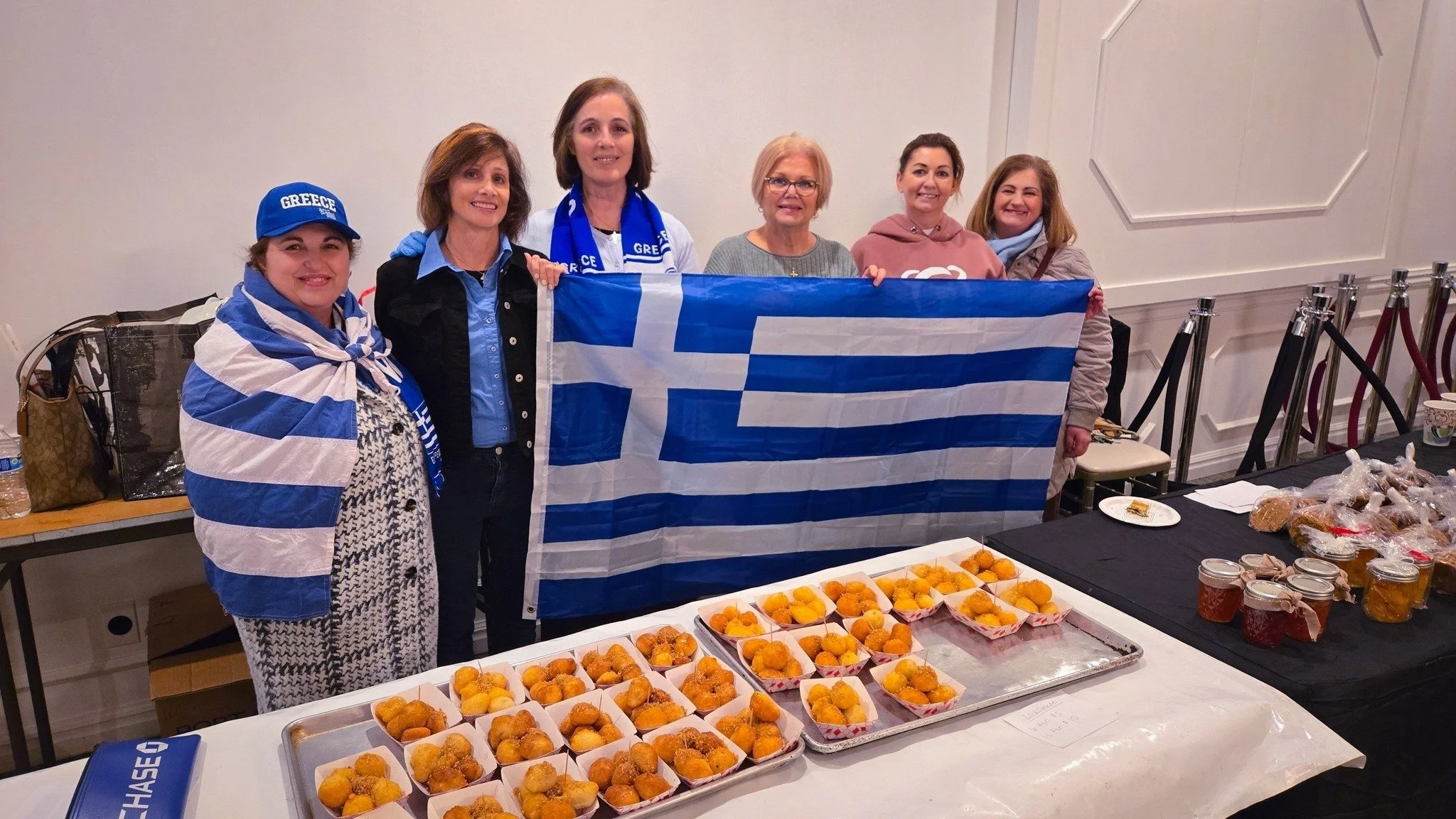 Group of six women standing behind a table with trays of food, holding a Greek flag, at what appears to be a cultural or community event.