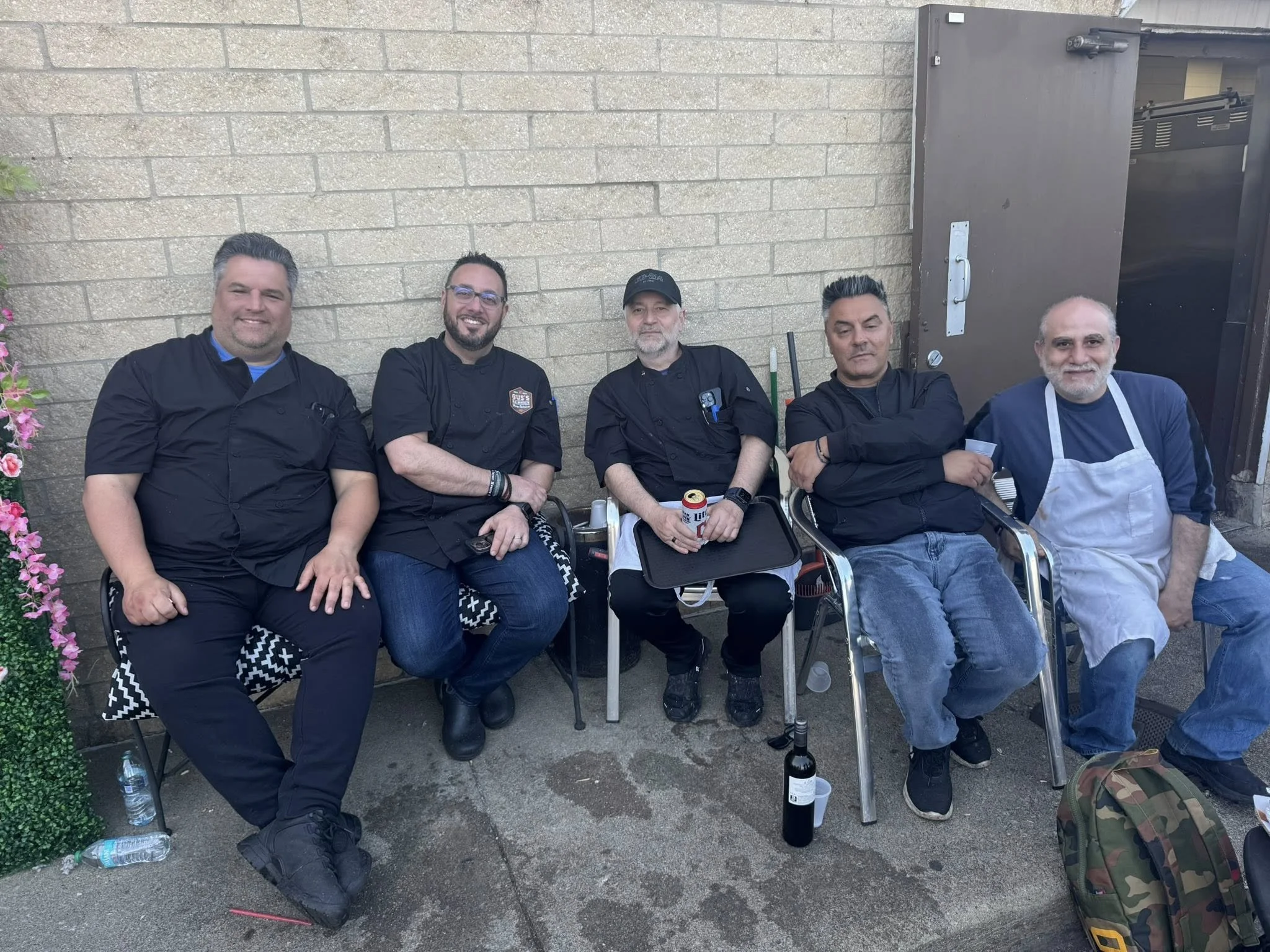 Five men sitting outdoors against a brick wall, some wearing kitchen uniforms, with drinks and a backpack near them.