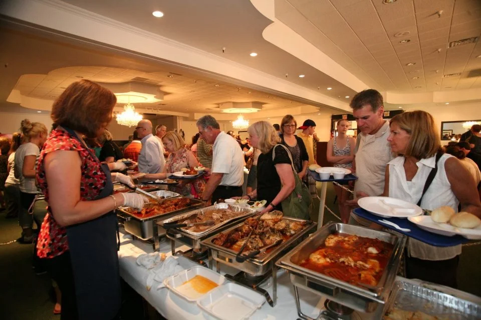 People serving themselves food from a buffet in a large room with ornate chandeliers and ceiling decorations.