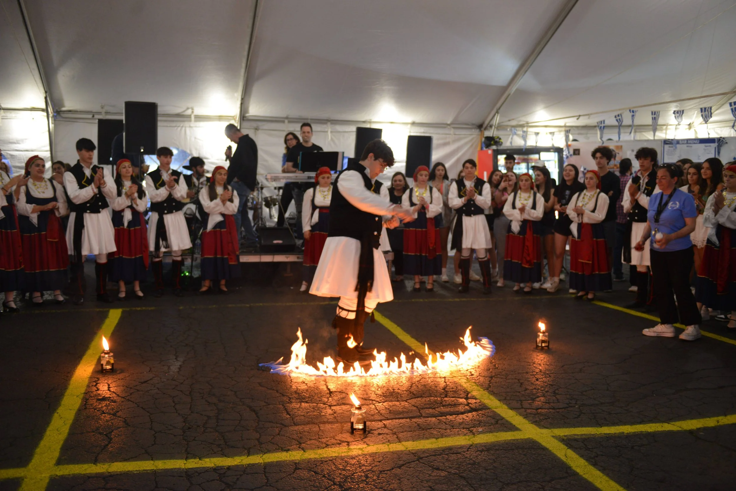 A man in traditional Greek clothing performing a fire dance in front of a group of people dressed in folk costumes at an indoor event.