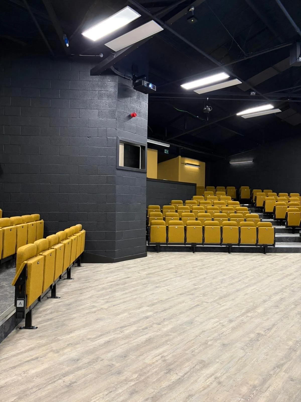 An empty lecture hall with yellow seats arranged in rows. The walls are painted black with a brick texture, and the ceiling has fluorescent lights. There is a small window on the left side of the wall.