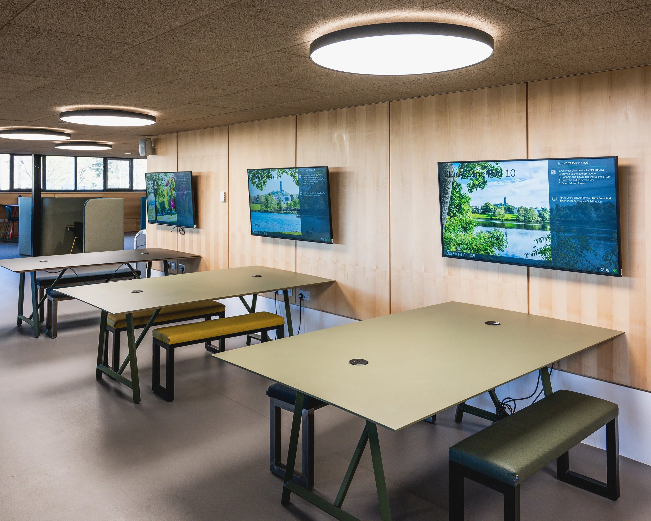 A conference room with multiple tables and chairs, several wall-mounted screens displaying landscape visuals and instructions, and ceiling lights.