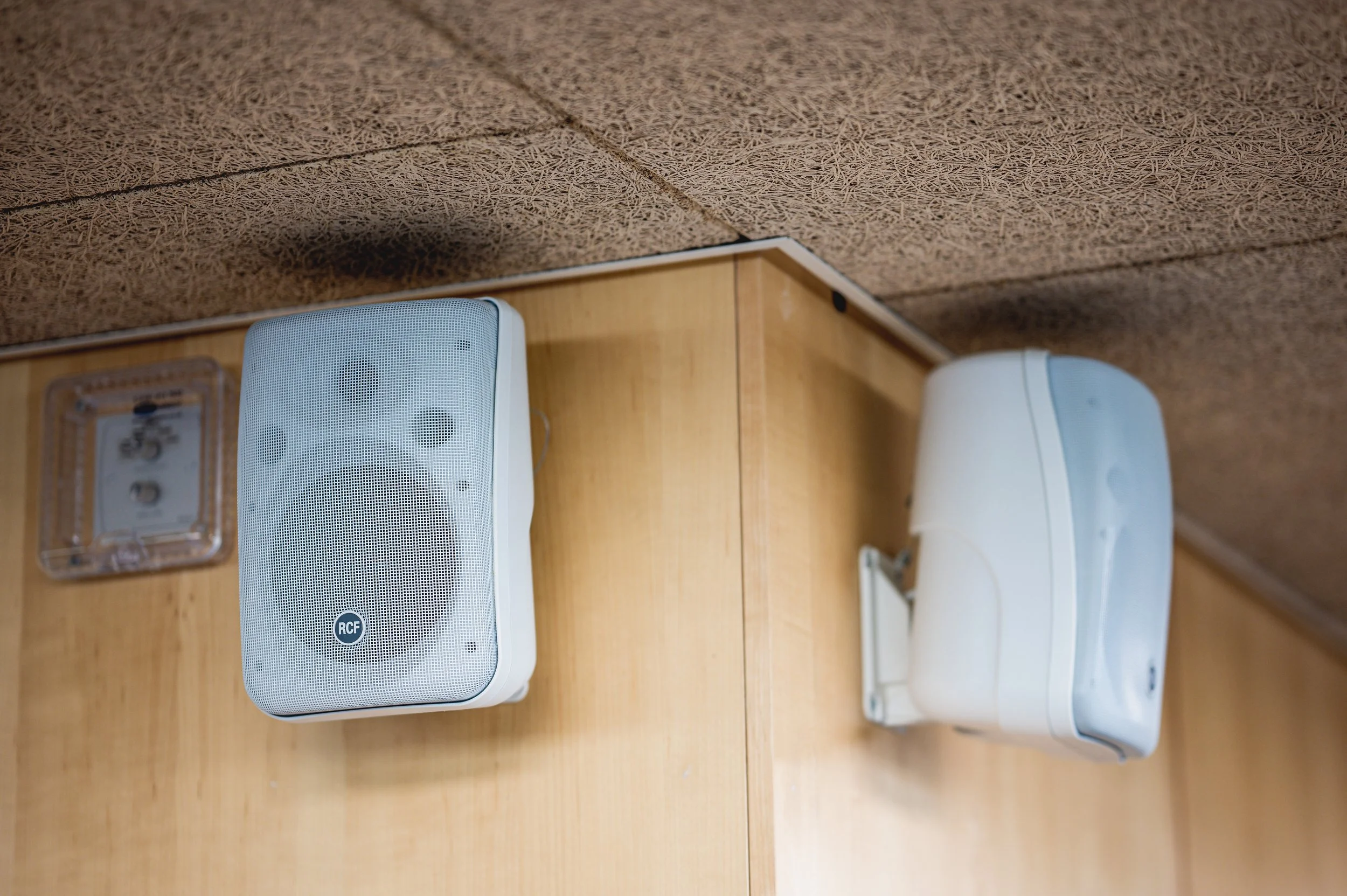 Two white rectangular speakers with rounded edges mounted on a wooden wall beneath a textured brown ceiling.