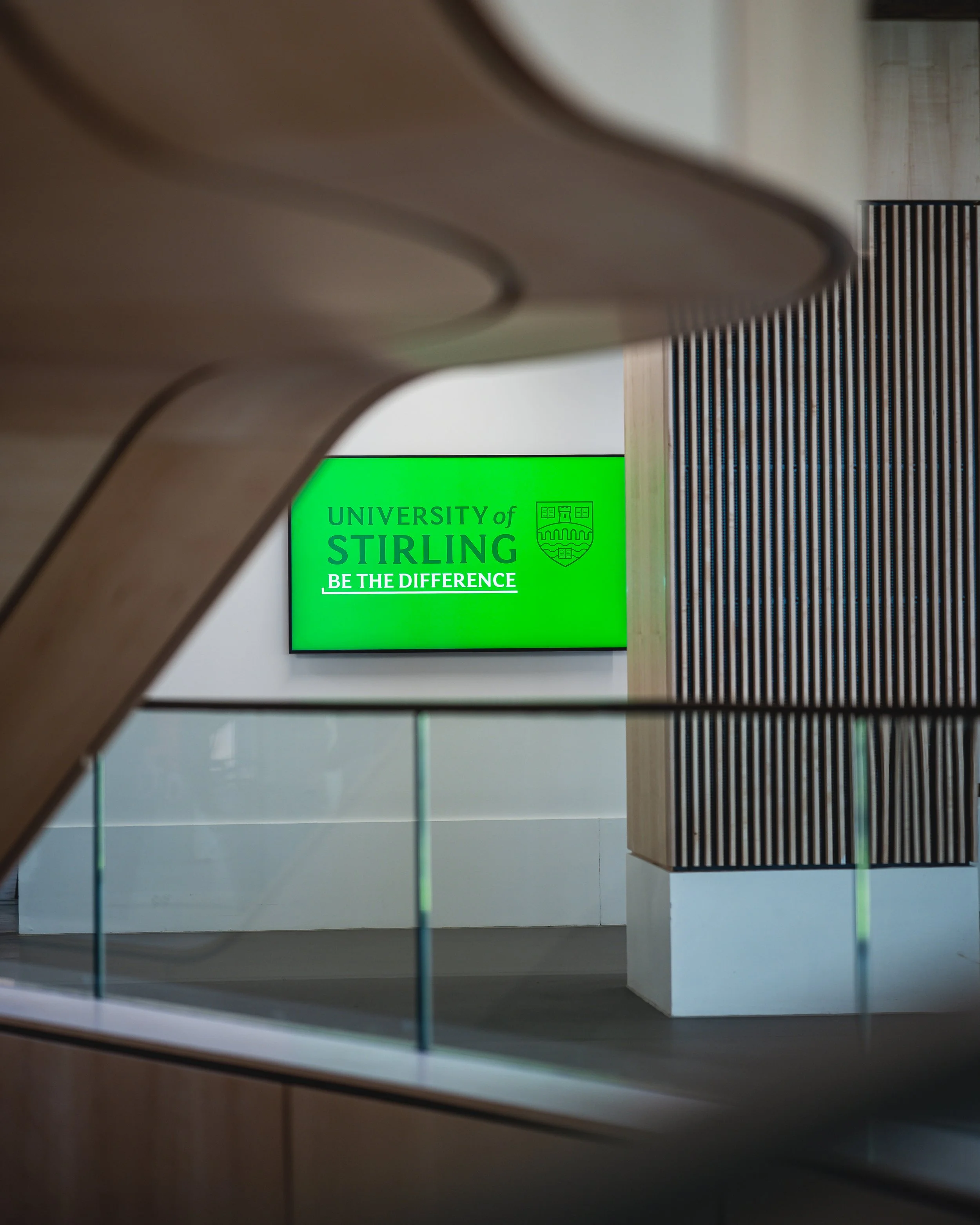 A green digital sign displaying the text 'University of Stirling Be the Difference' with a white background, viewed through a wooden and glass railing.