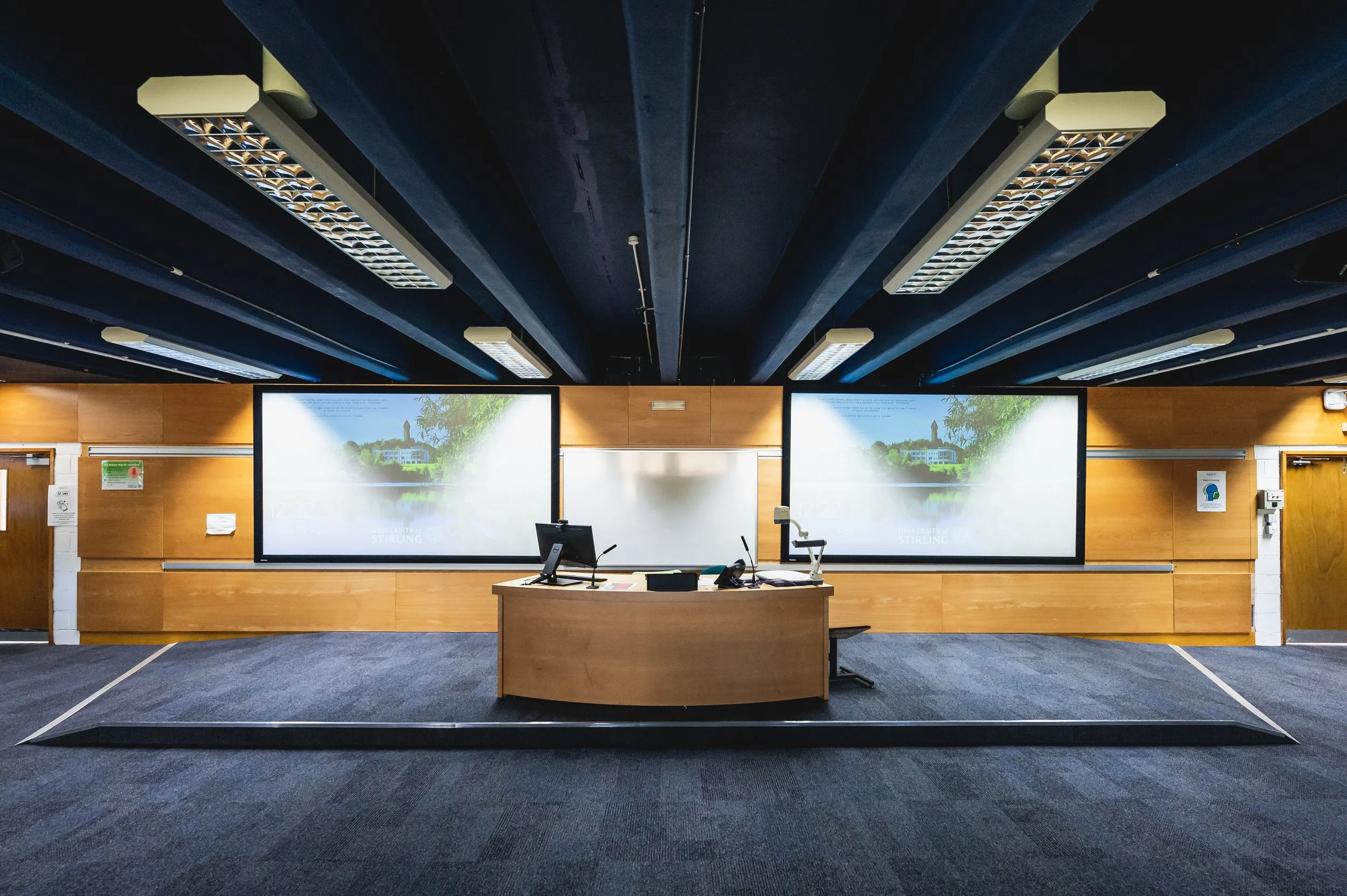 An empty classroom or seminar room with two large presentation screens displaying a scenic view of trees and a building, a desk with a computer, microphone, and documents, and overhead fluorescent lights on a dark ceiling.