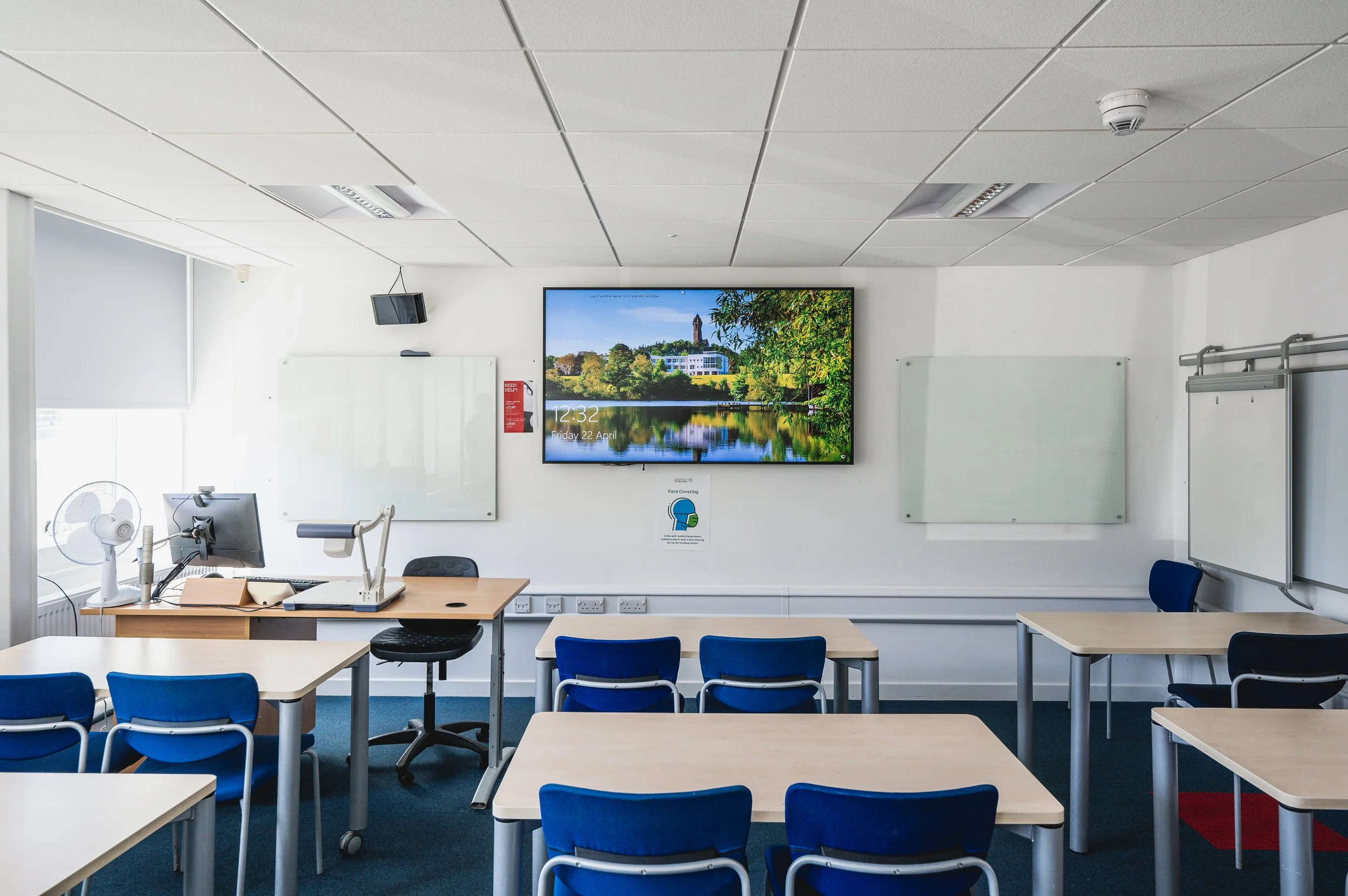 A classroom with white walls, a large wall-mounted screen displaying a scenic view of a lake and trees, whiteboards, desks with blue chairs, a computer on a desk, and a ceiling with fluorescent lights.