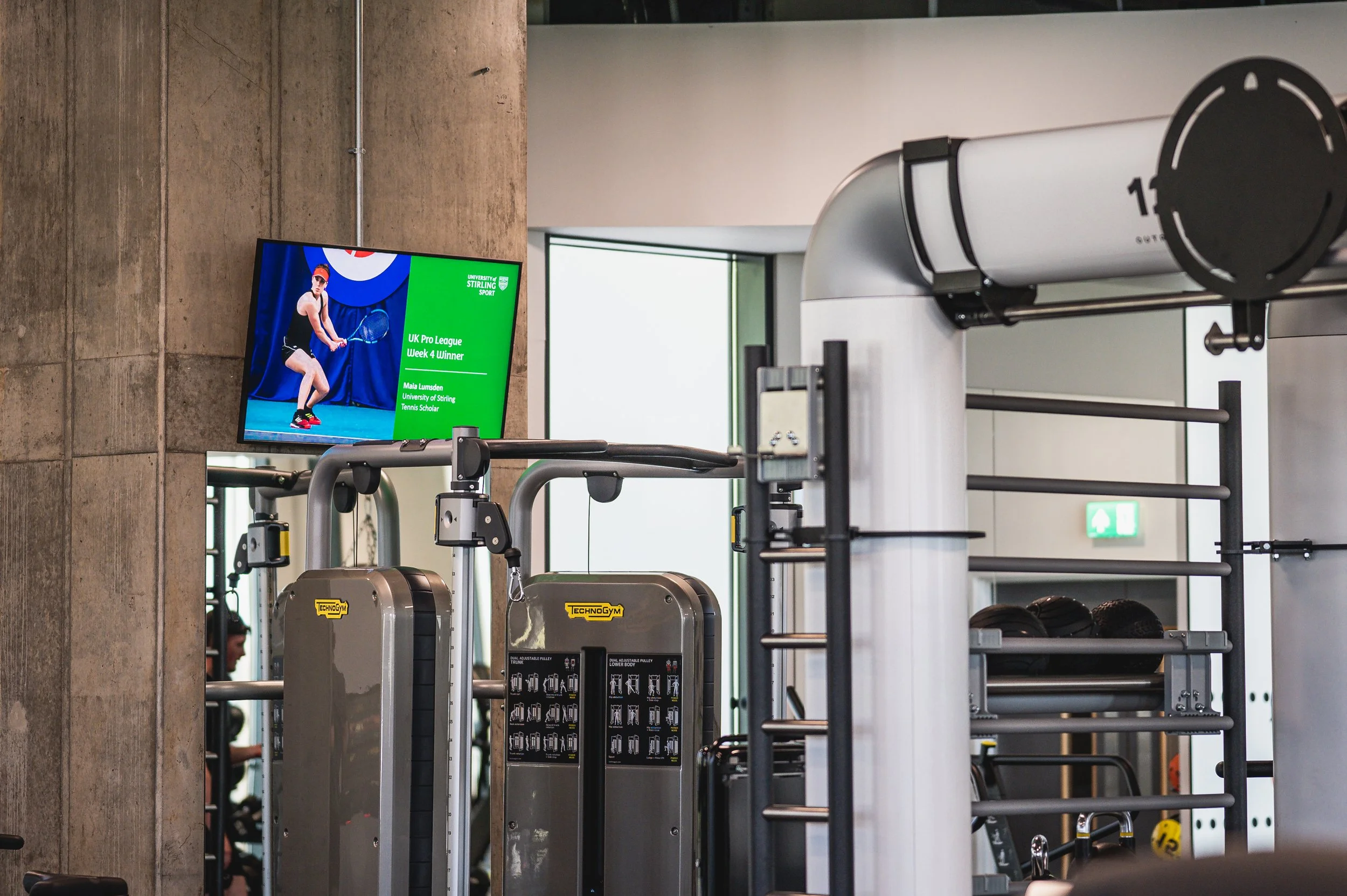 Interior of a gym with workout machines, a wall-mounted screen displaying sports news, and windows letting in natural light.
