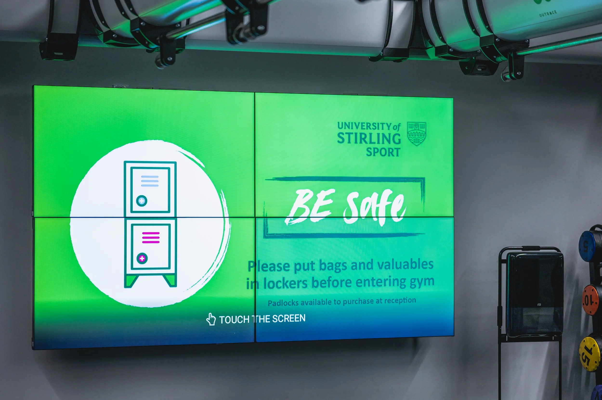Digital signage at the University of Stirling Sport with safety instructions, including reminders to put bags and valuables in lockers before entering the gym, and available padlocks for purchase at reception.