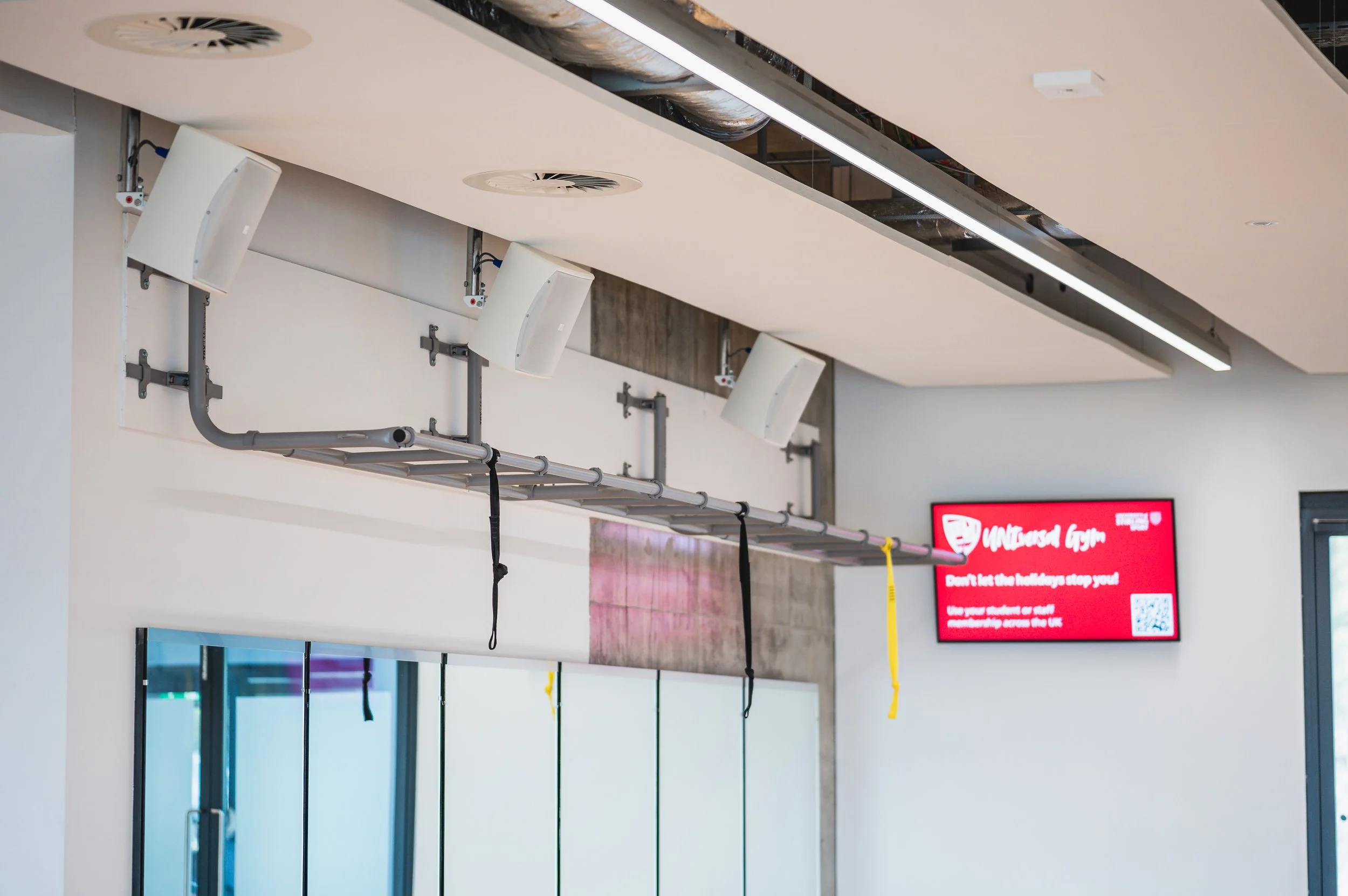 Interior of a gym with white walls and ceiling, displaying a row of white speakers, black pipes with hanging straps, ventilation ducts, and a digital screen in the background.