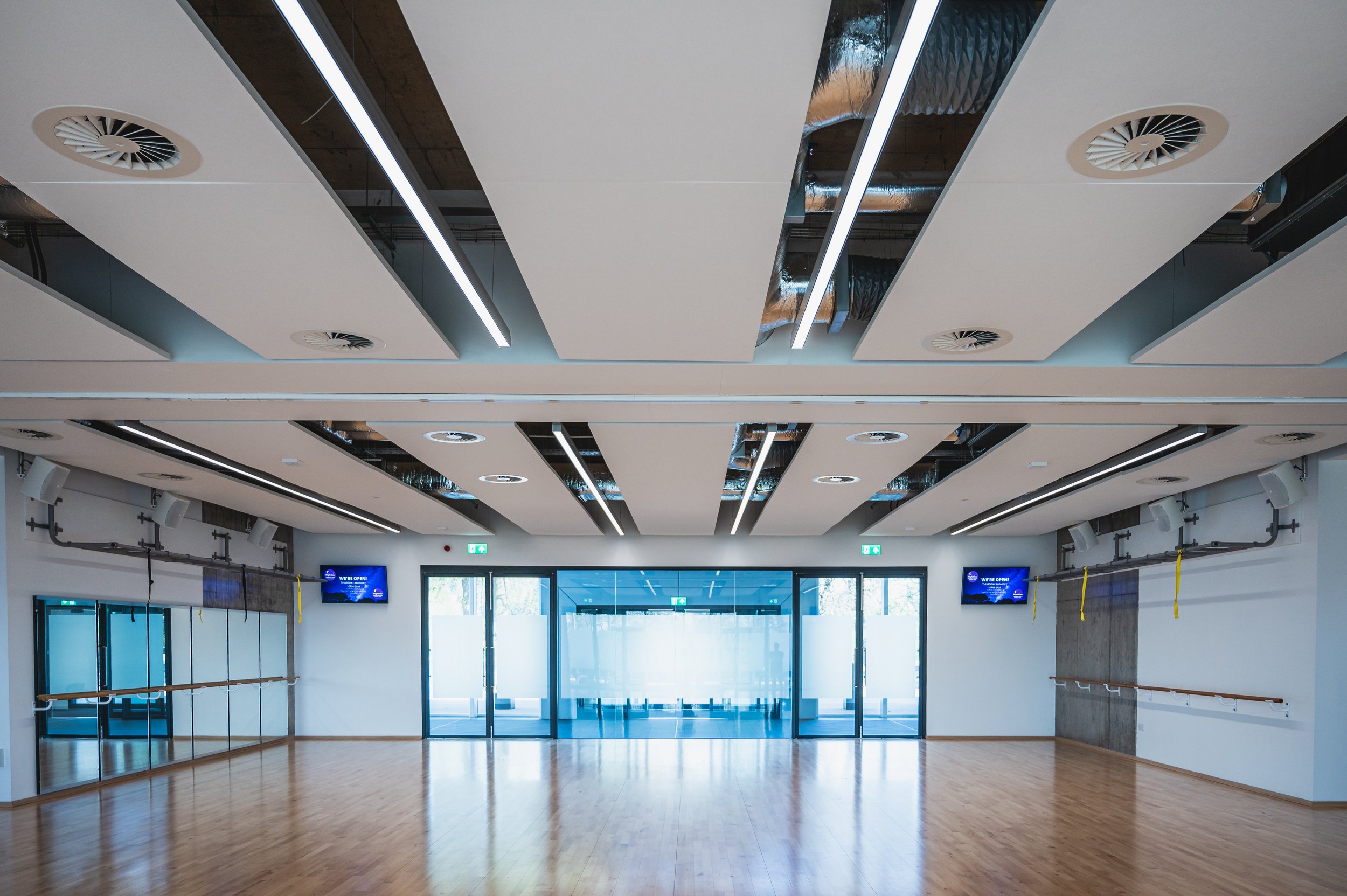 Interior view of a modern dance or fitness studio with wooden floors, mirrors, ballet bars, and a high ceiling with vents and long lighting fixtures, near glass doors.