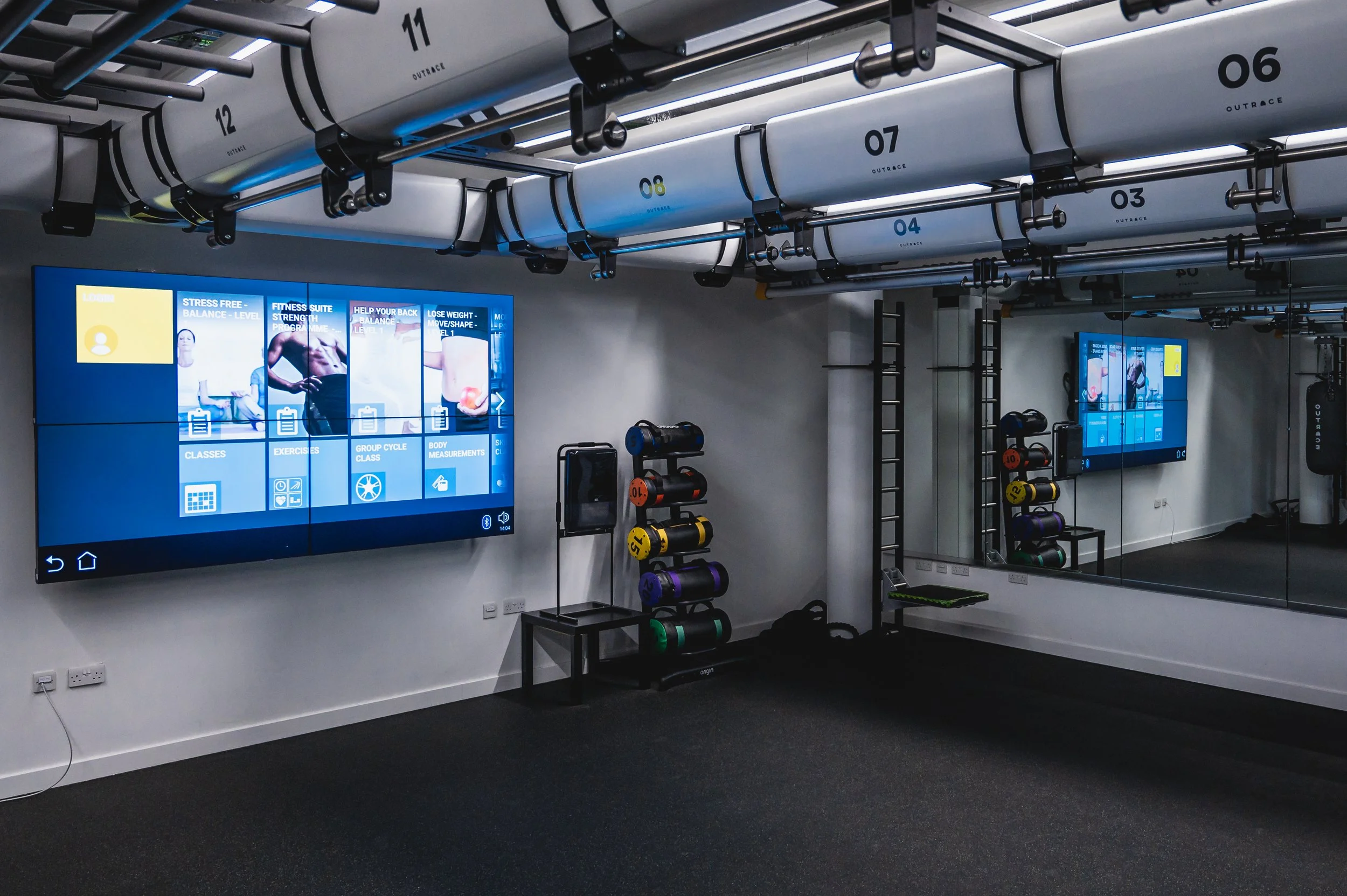 Interior of a modern gym with concrete pipes labeled with numbers and the word 'Outrace' suspended from the ceiling, exercise equipment, and digital screens displaying fitness information
